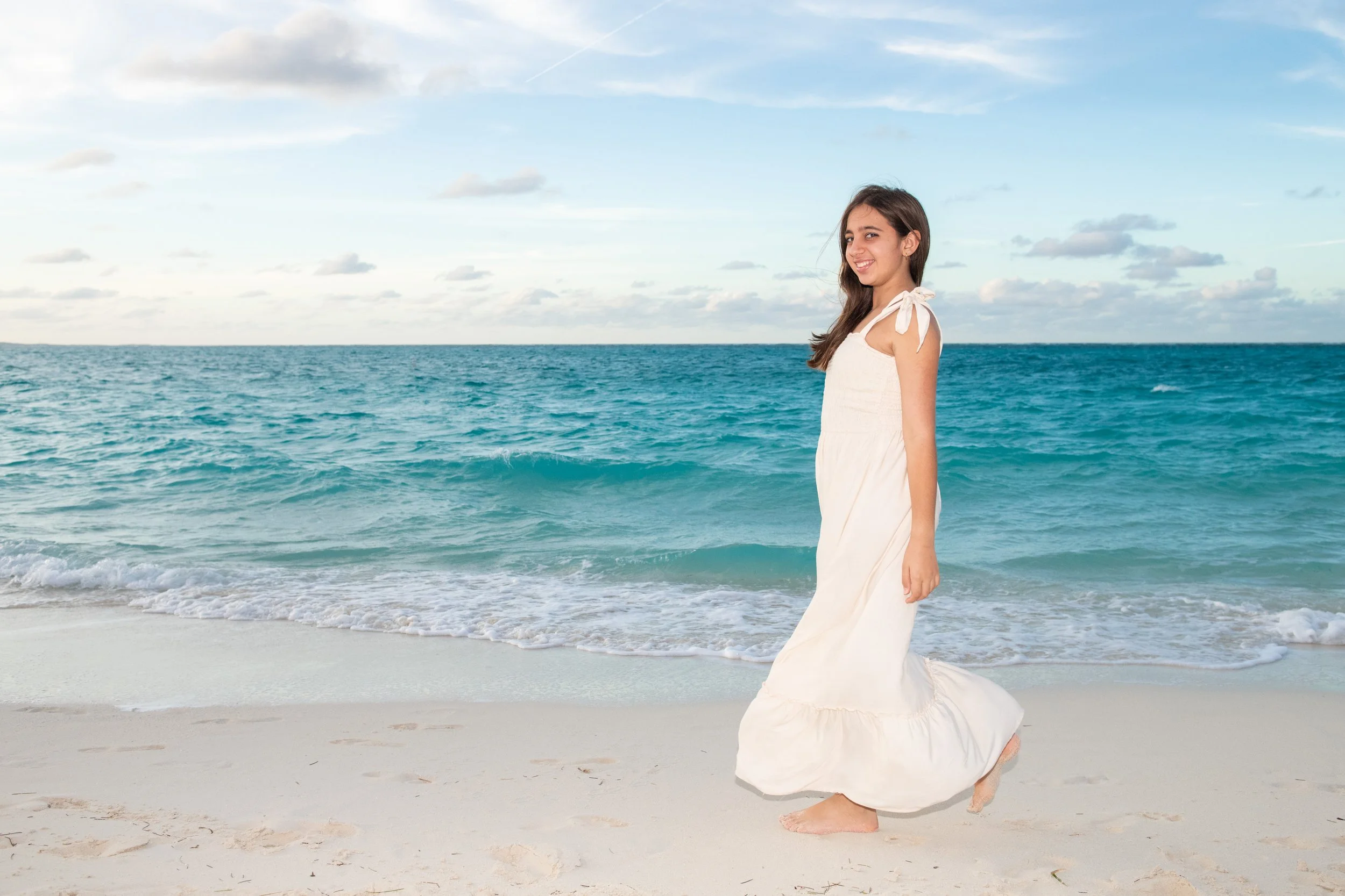 Layla Kazerouni, a young woman in a white dress walking barefoot on a sandy beach near the ocean, with a partly cloudy sky overhead.