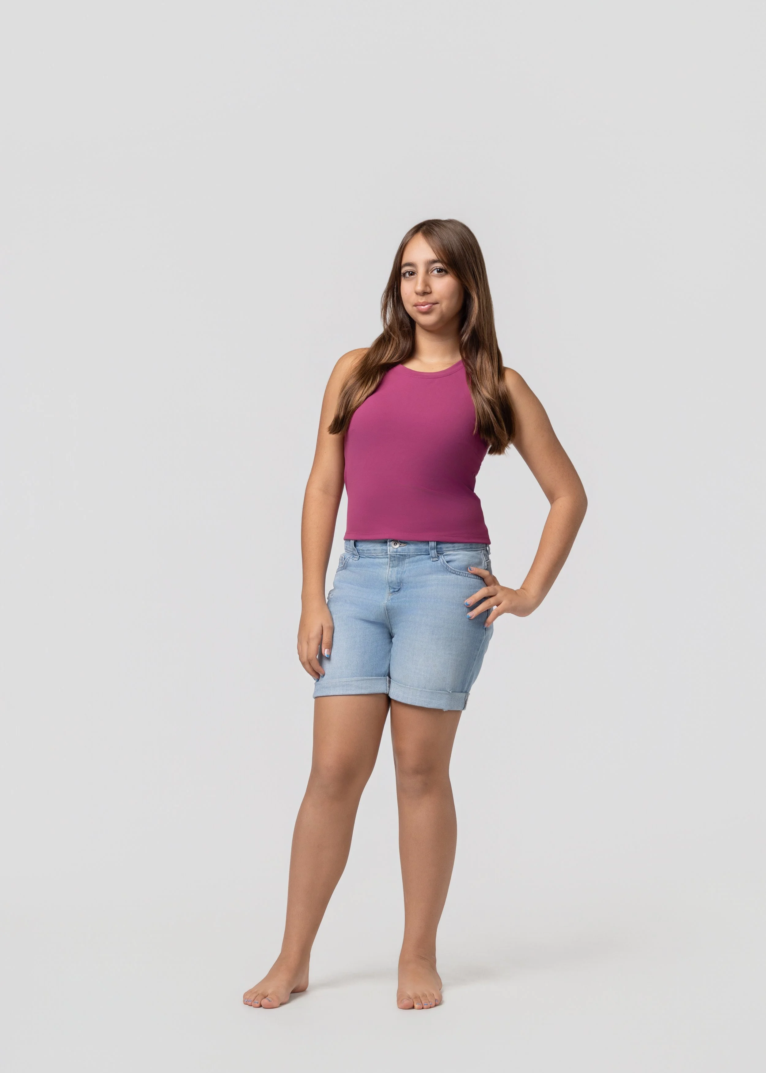 Layla Kazerouni, a young woman standing barefoot against a plain white background. She is wearing a sleeveless purple top and light blue denim shorts, with her right hand resting on her hip and her left arm hanging loosely by her side.