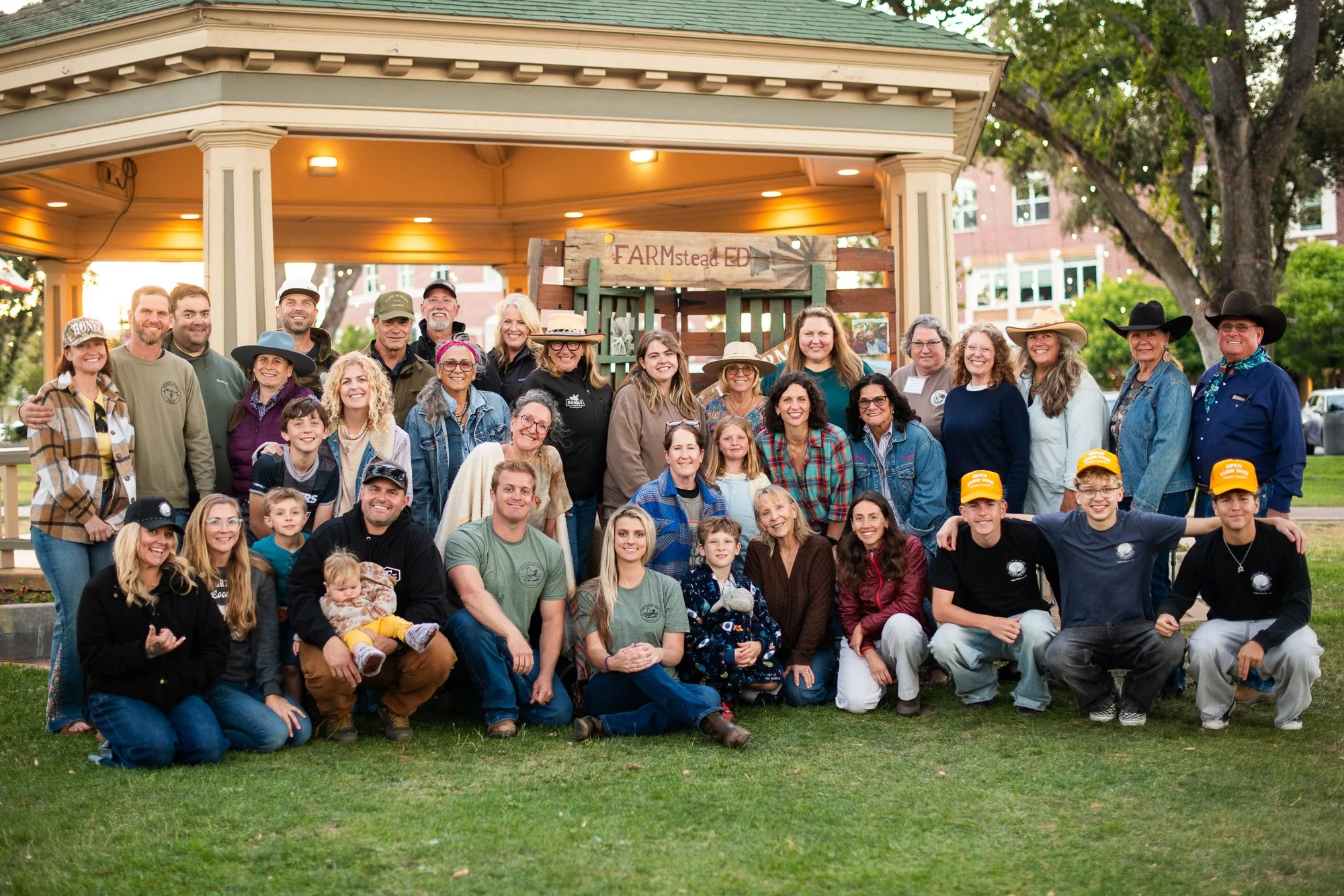 A large group of people, including children, adults, and seniors, gathered outdoors in front of a gazebo and a wooden sign that reads 'FARMstead ED,' during a community event in a park on a clear day with trees and buildings in the background.