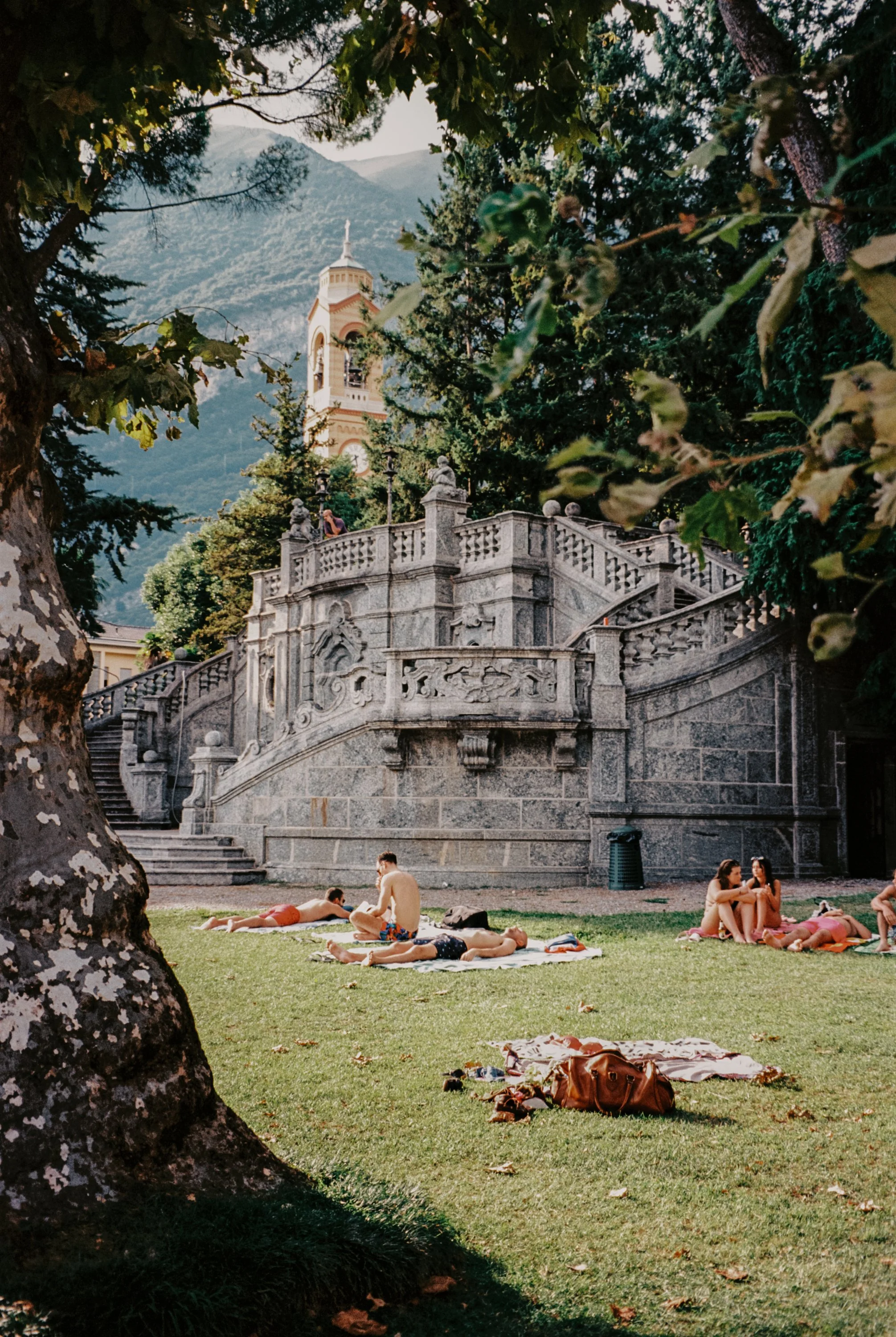 People relaxing and sunbathing on towels on a grassy area, with a historic stone staircase and a church tower with mountains in the background, shaded by trees. Travel places photography by Lenneke Benders. 