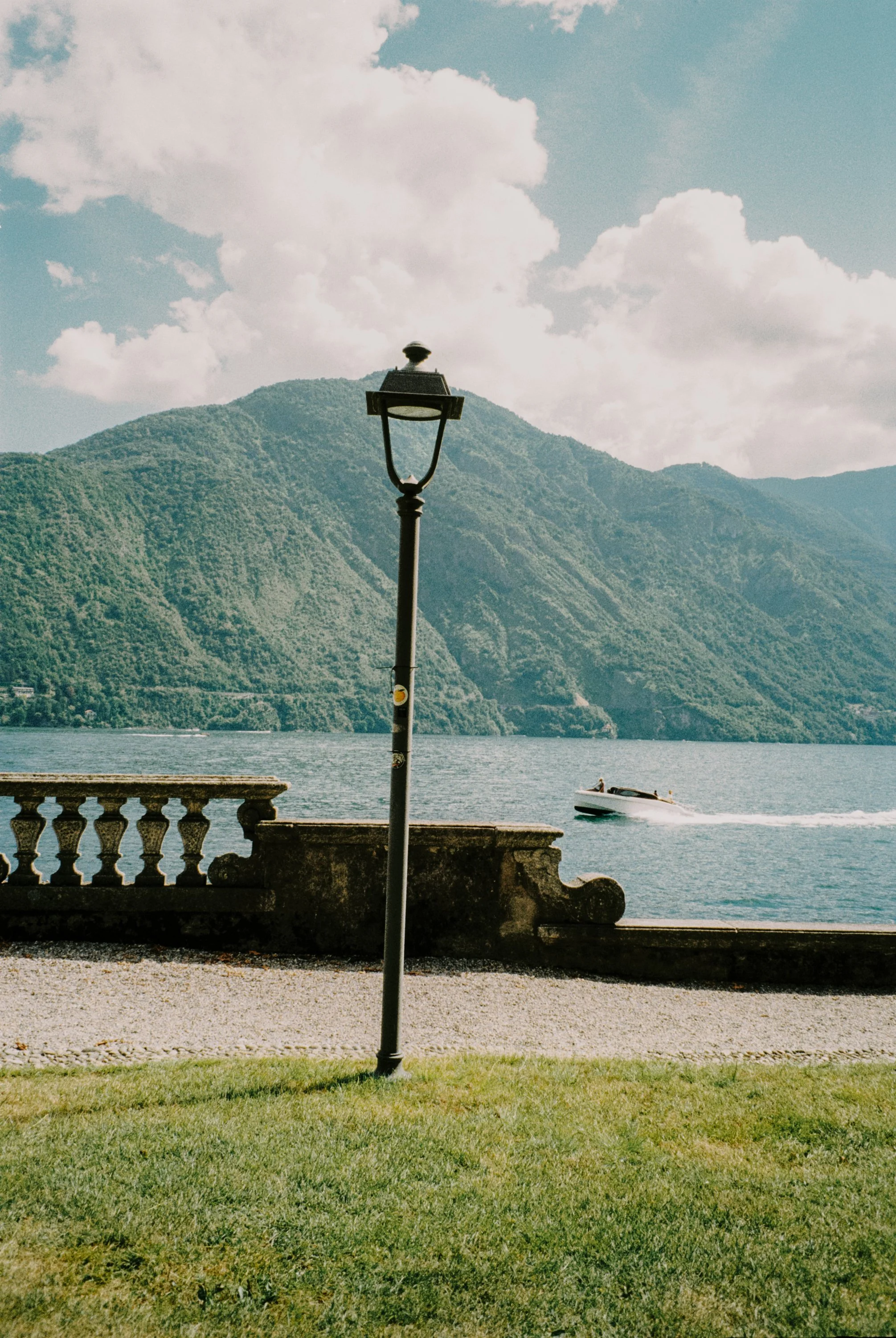 A lakeside scene with a stone railing, a street lamp, a grassy area in the foreground, water, a boat in motion, and green mountains in the background under partly cloudy sky. Fine art photography by Lenneke Benders in Como. 