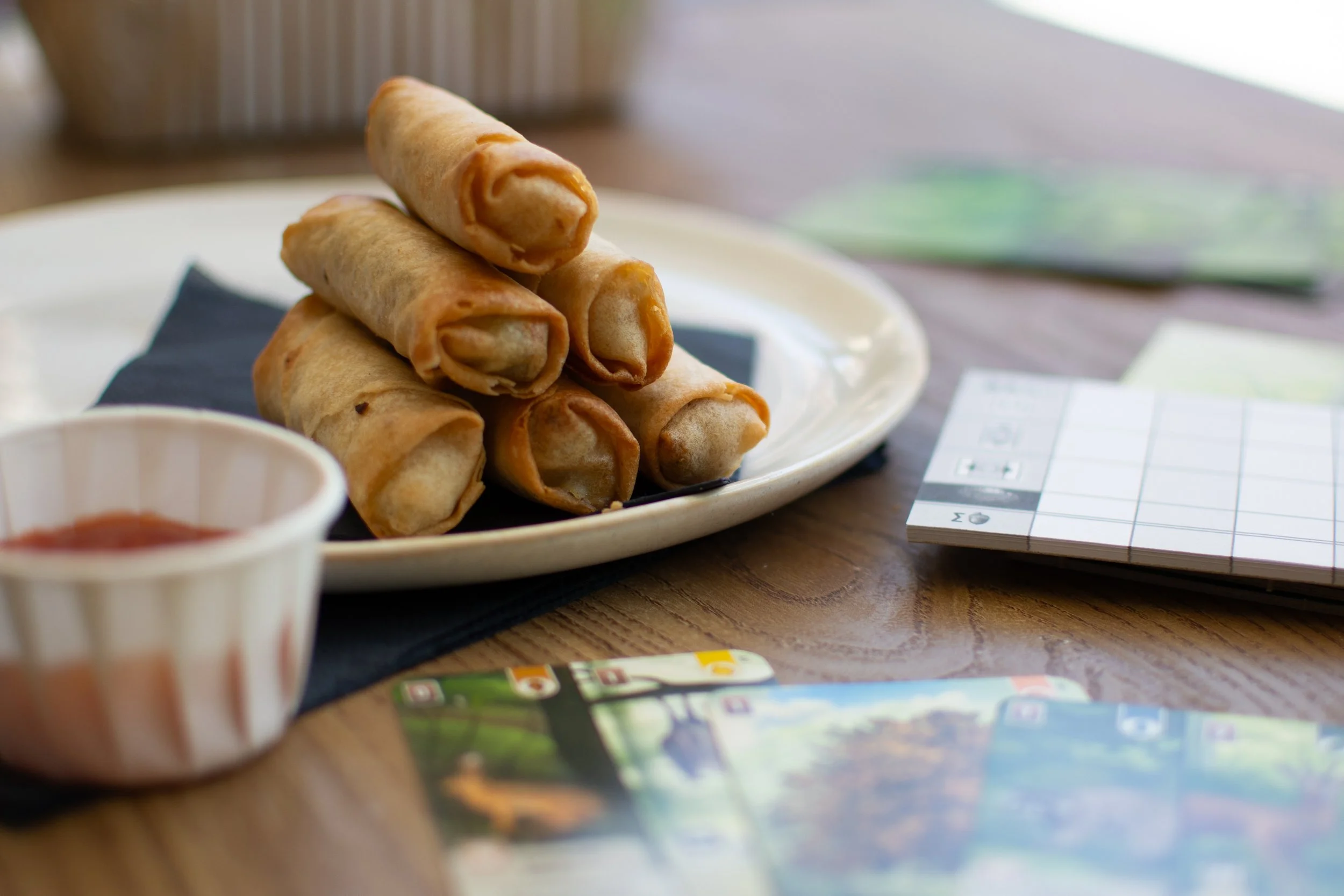 Close-up of a plate with homemade spring rolls on a black napkin, accompanied by a cup of sweet chili sauce, with colorful brochures and a calendar on a wooden table in the background.