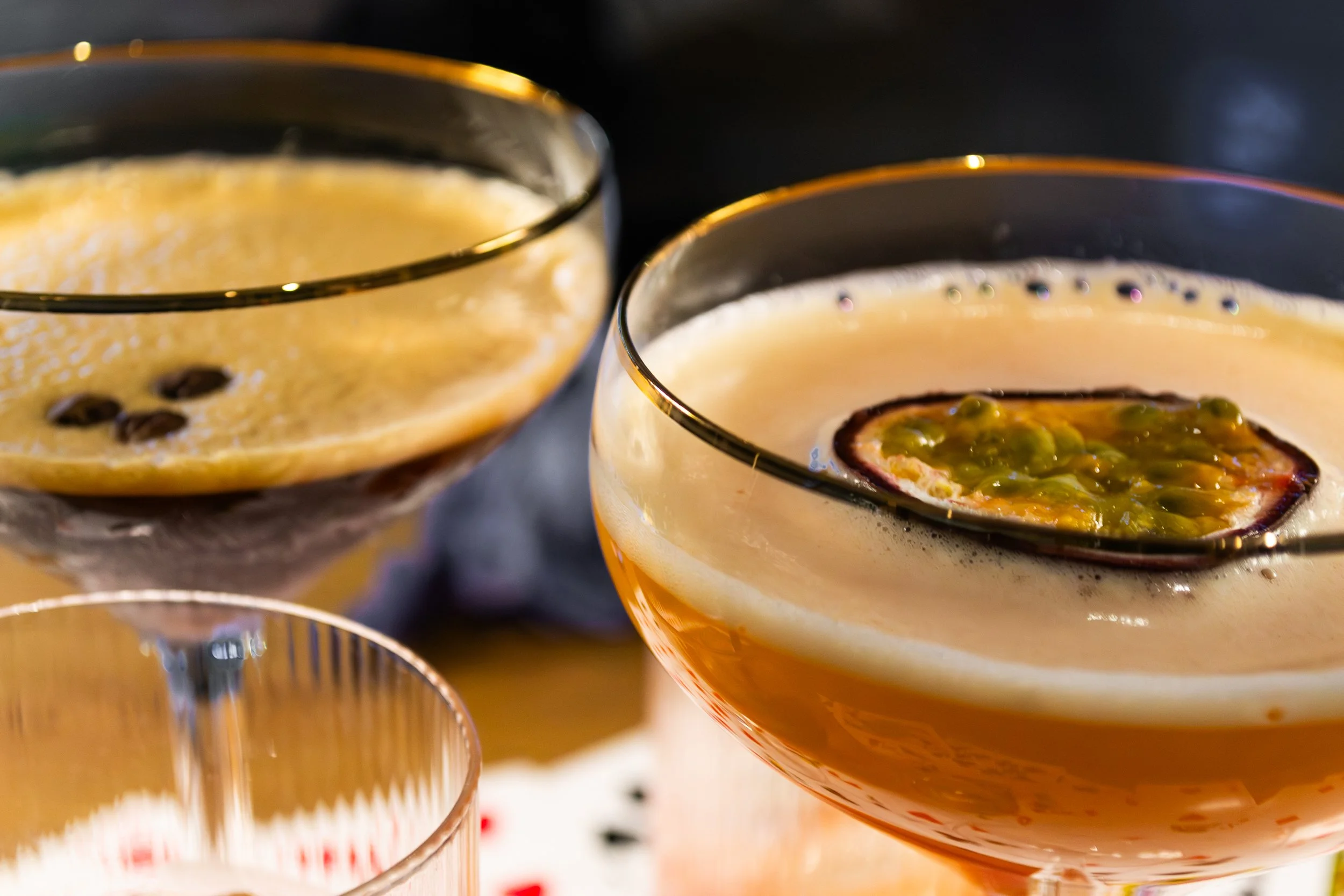Close-up of two glasses filled with a creamy cocktail, one garnished with passion fruit and the other with coffee beans, set against a dark background.