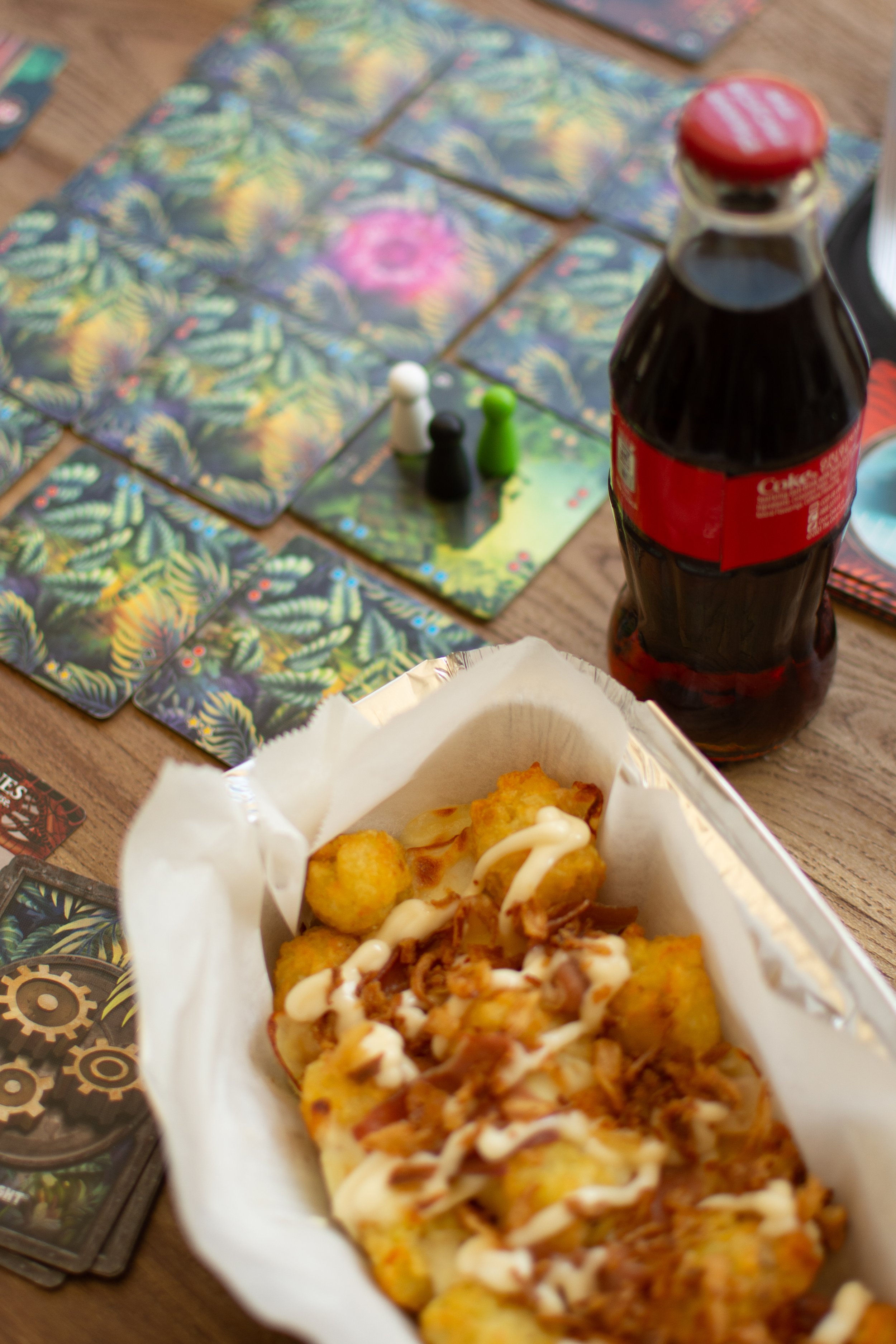 Close-up of a serving of potato tots topped with bacon bits and drizzled with mayonnaise and barbecue sauce in a paper food container. In the background, there are colorful game cards and a bottle of Coca-Cola on a wooden table.
