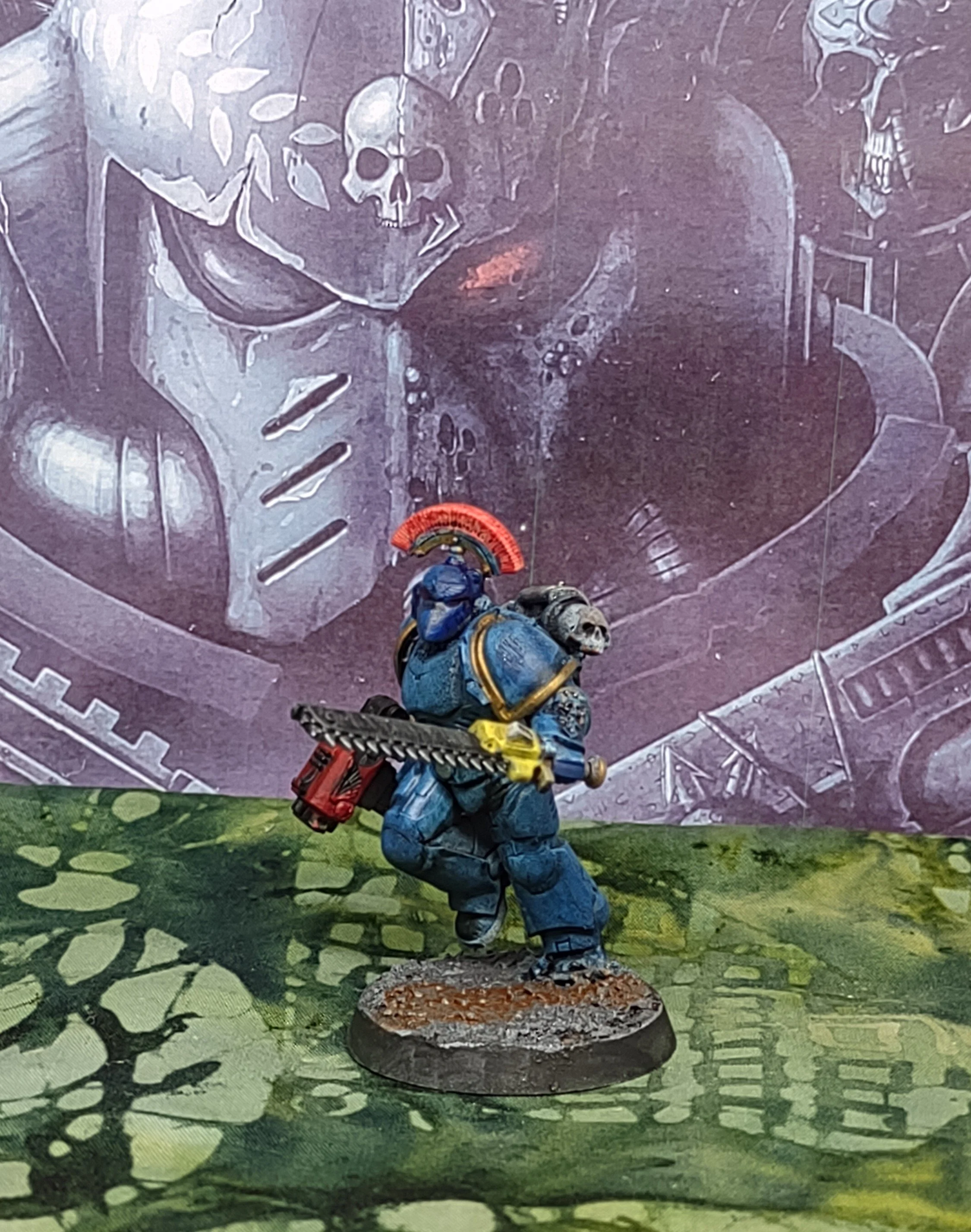 Miniature figurine of a sci-fi soldier with a skull on the shoulder pad, holding a chainsaw, wearing blue armor with a red helmet, positioned on a home made base with a painted backdrop featuring a skull and mechanical elements.
