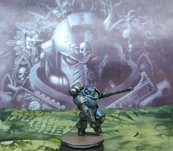 A miniature figurine of a futuristic soldier with a gun and sword, standing on a terrain base in front of a painted backdrop depicting a mechanical skull with a horn, surrounded by skulls and machinery.