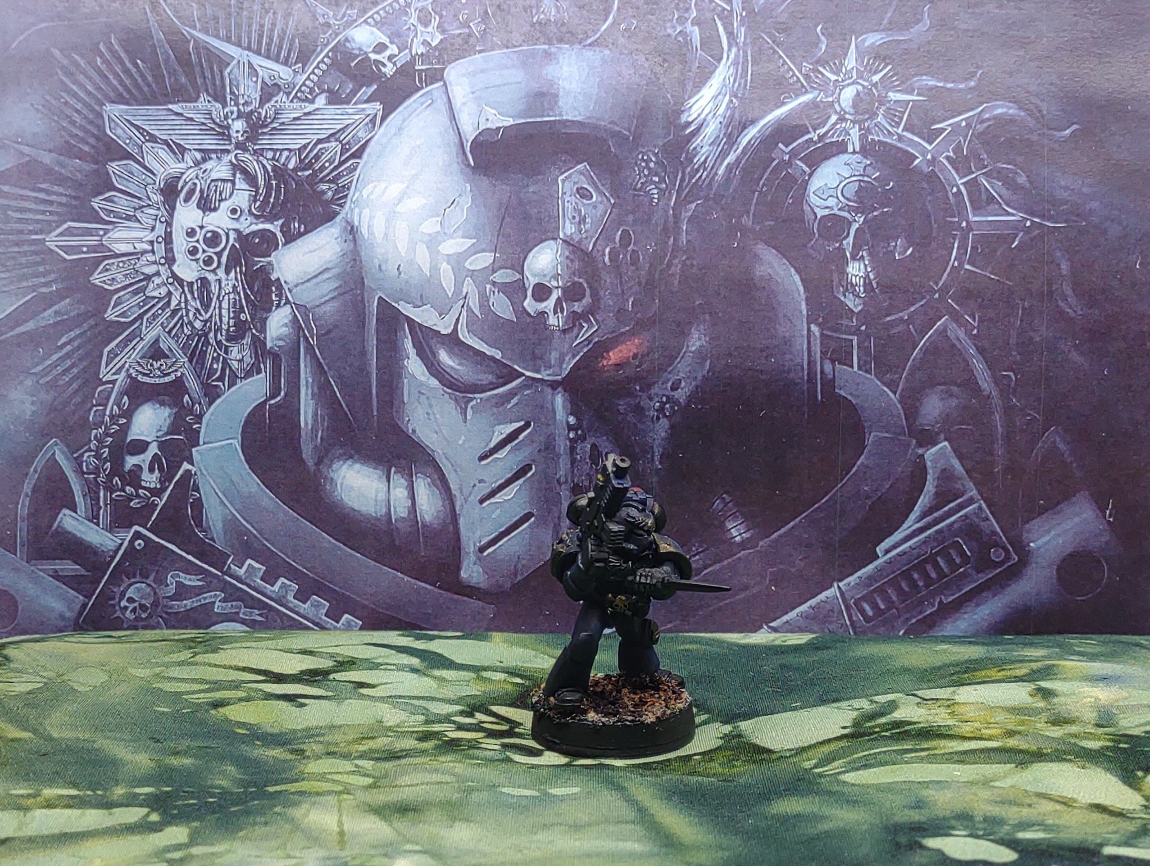 Miniature figurine of a soldier or space marine holding a weapon, standing on a textured base with a green landscape backdrop. In the background, there is a large wall mural depicting a sci-fi or fantasy scene with skulls, a helmet, and mechanical or alien elements.