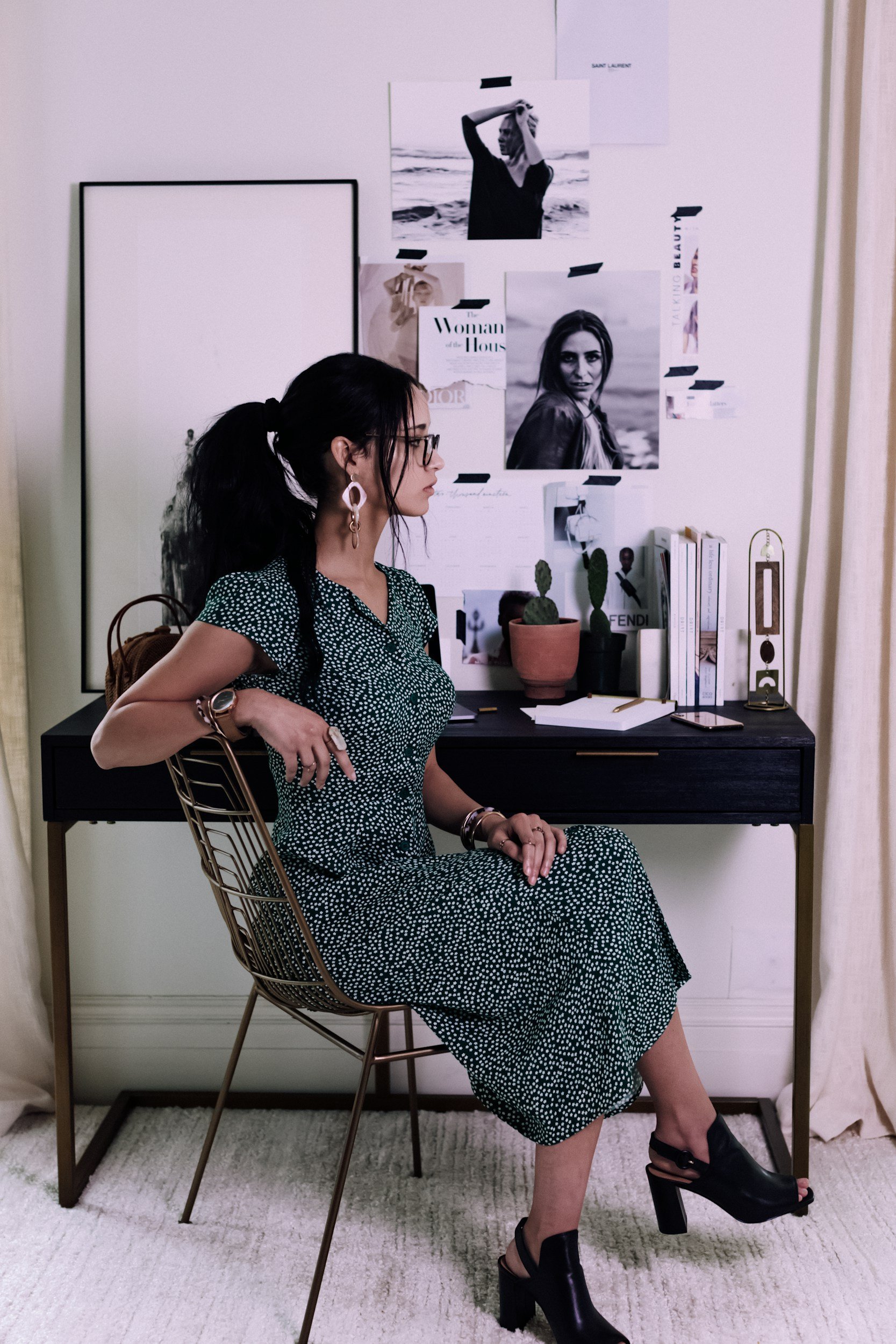 A woman sitting sideways in a chair, wearing a green polka dot dress, black high-heels, and glasses, with black hair in braids, in front of a white wall decorated with black-and-white photographs and magazines.