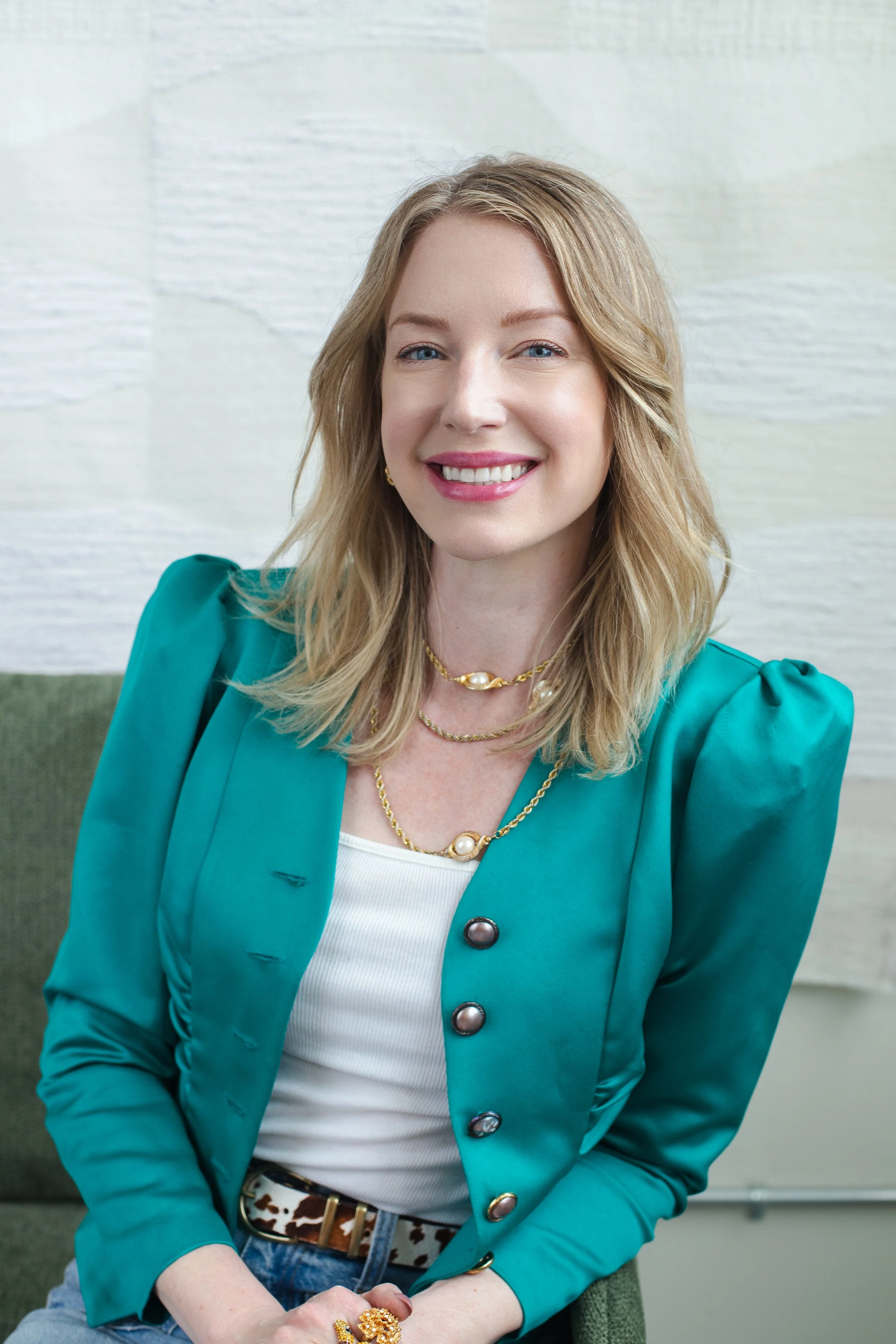 A woman with blonde hair, blue eyes, and a bright smile, wearing a turquoise blazer with large buttons, layered gold necklaces, a white top, and a leopard-print belt, seated against a light-colored textured wall.