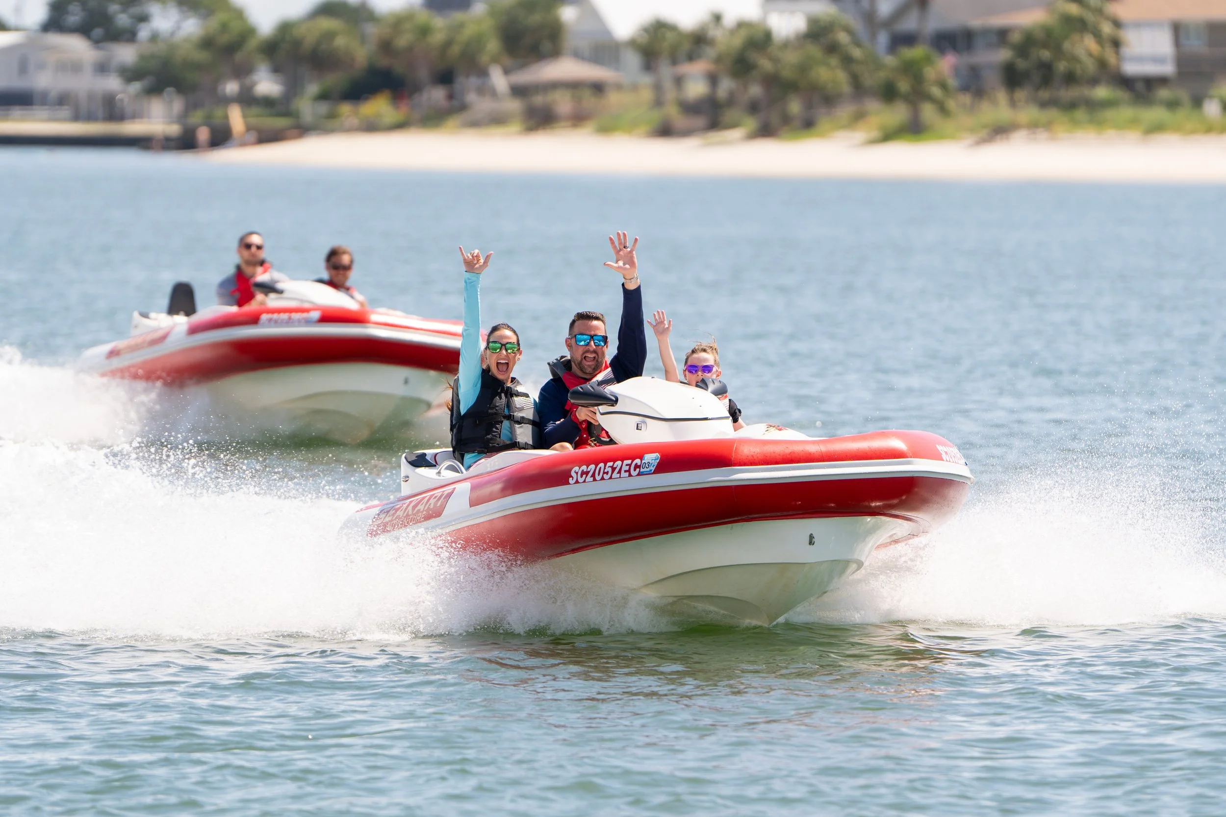 Group of people riding on red and white Seakarts, waving and smiling, on a body of water with shoreline and buildings in the background.