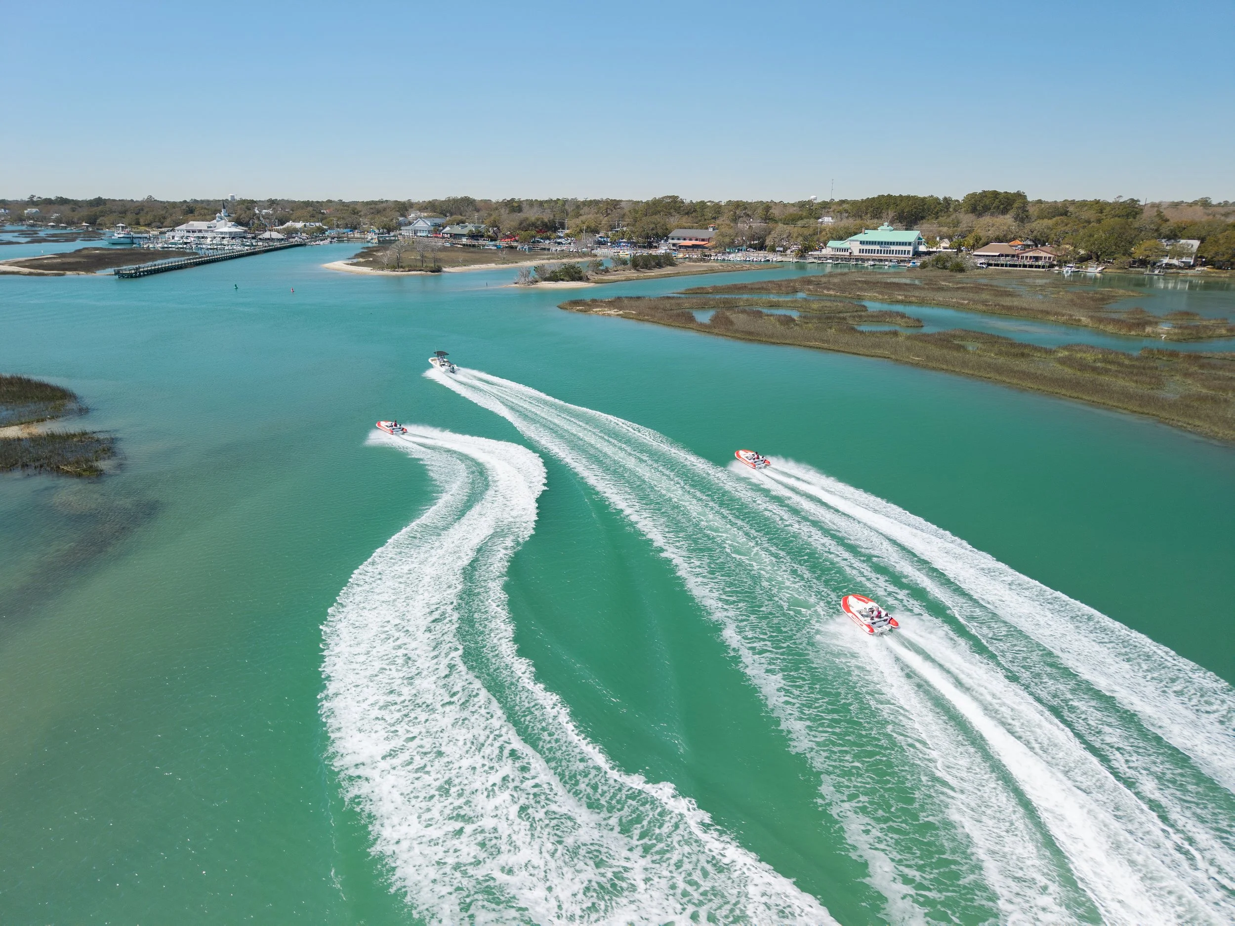 Four Seakarts speeding across Murrells Inlet, SC, creating white wake trails, with The Marsh Walk in the background under a clear blue sky.