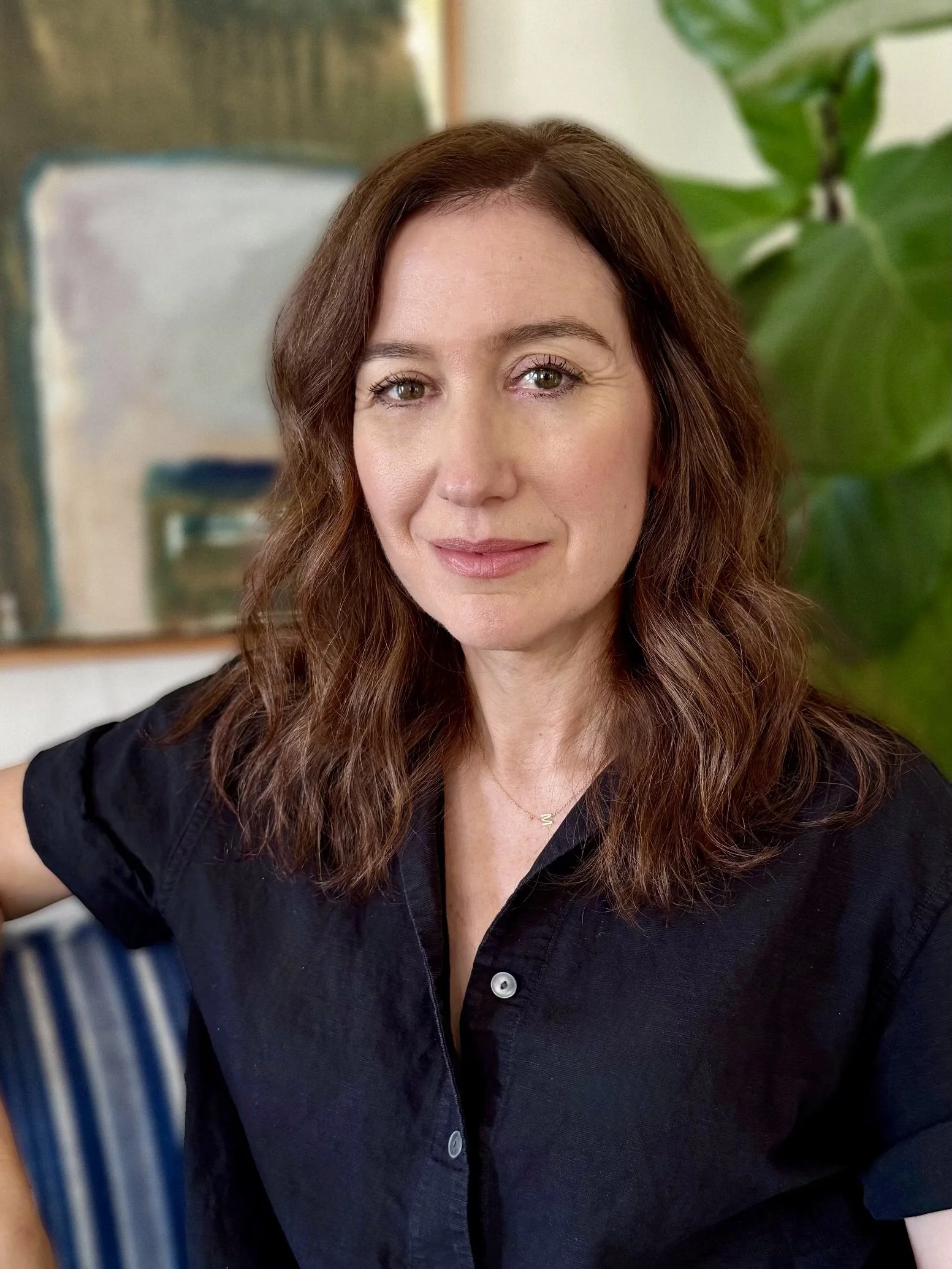 A woman with shoulder-length wavy brown hair, wearing a black button-up shirt, sitting indoors with a large green plant and a blurred colorful painting in the background.