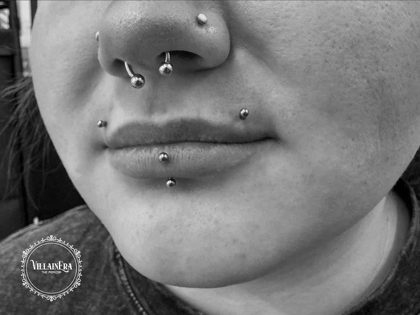 Close-up of a person's face showing multiple facial piercings, including nose, lip, and chin piercings, in black and white.