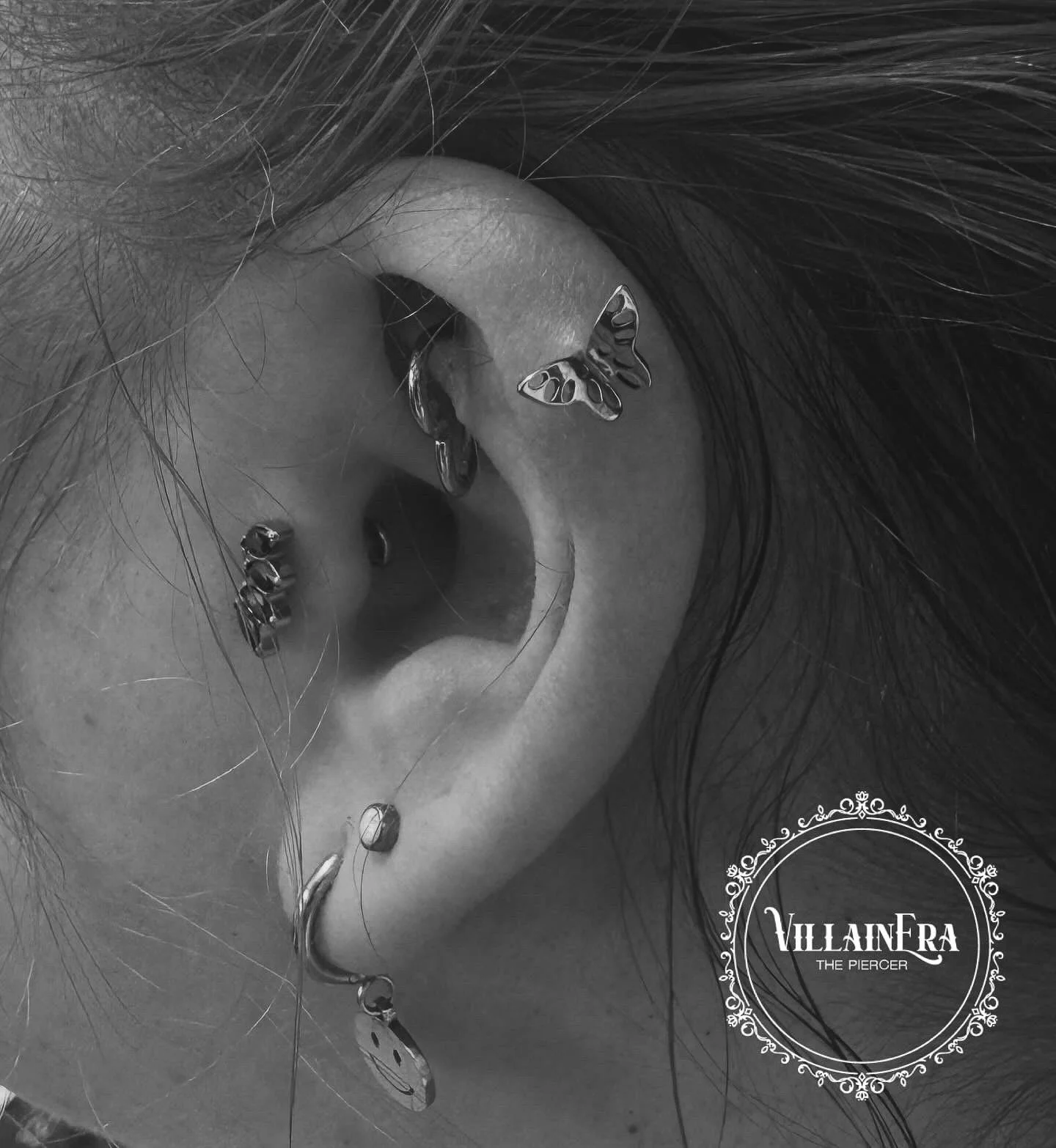 Close-up of a person's ear with multiple earrings, including a butterfly-shaped earring, a smiley face charm, and other small hoops, with dark hair and a decorative circular logo reading 'VILLAINFRA THE PIERCER' in the corner.