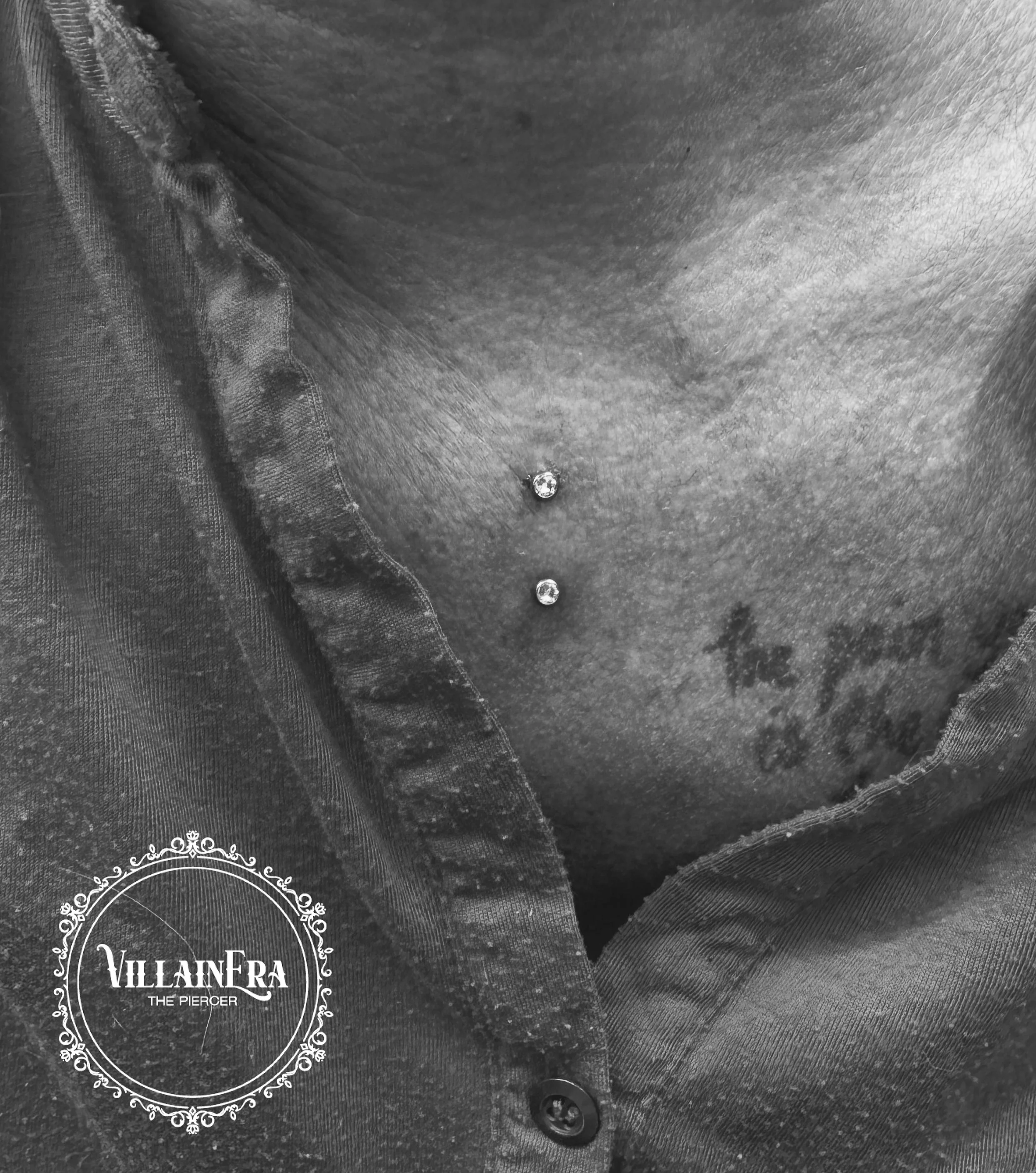 Close-up of a person's neck with two small diamond piercings and tattoos, partially covered by a torn shirt with a VillainEra logo in the lower left corner.