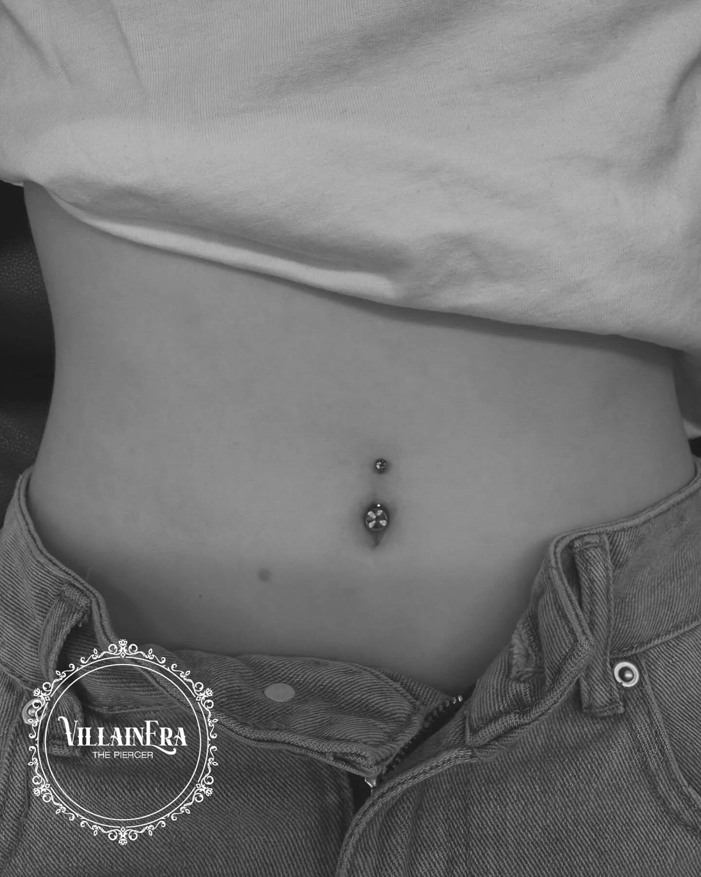 Close-up of a person's midsection showing a belly button piercing with a decorative gem, wearing jeans slightly unbuttoned, with a t-shirt pulled up to reveal the piercings.