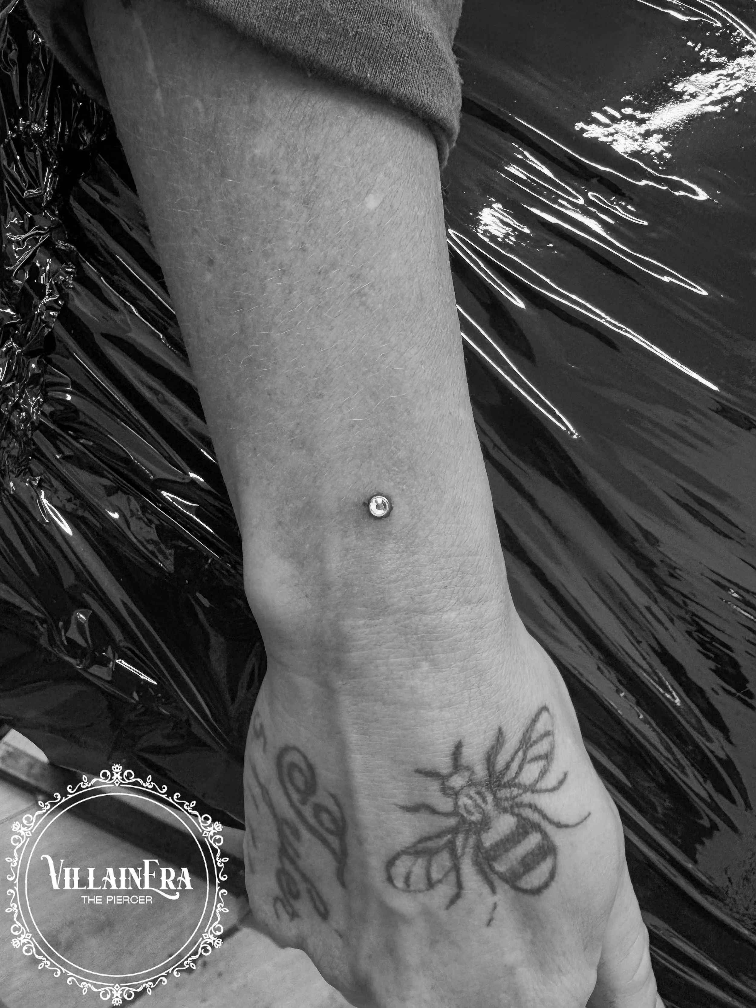 A person's forearm decorated with a bee tattoo and a script tattoo, with a fresh piercing on the wrist featuring a small gemstone, set against a background of shiny black material.