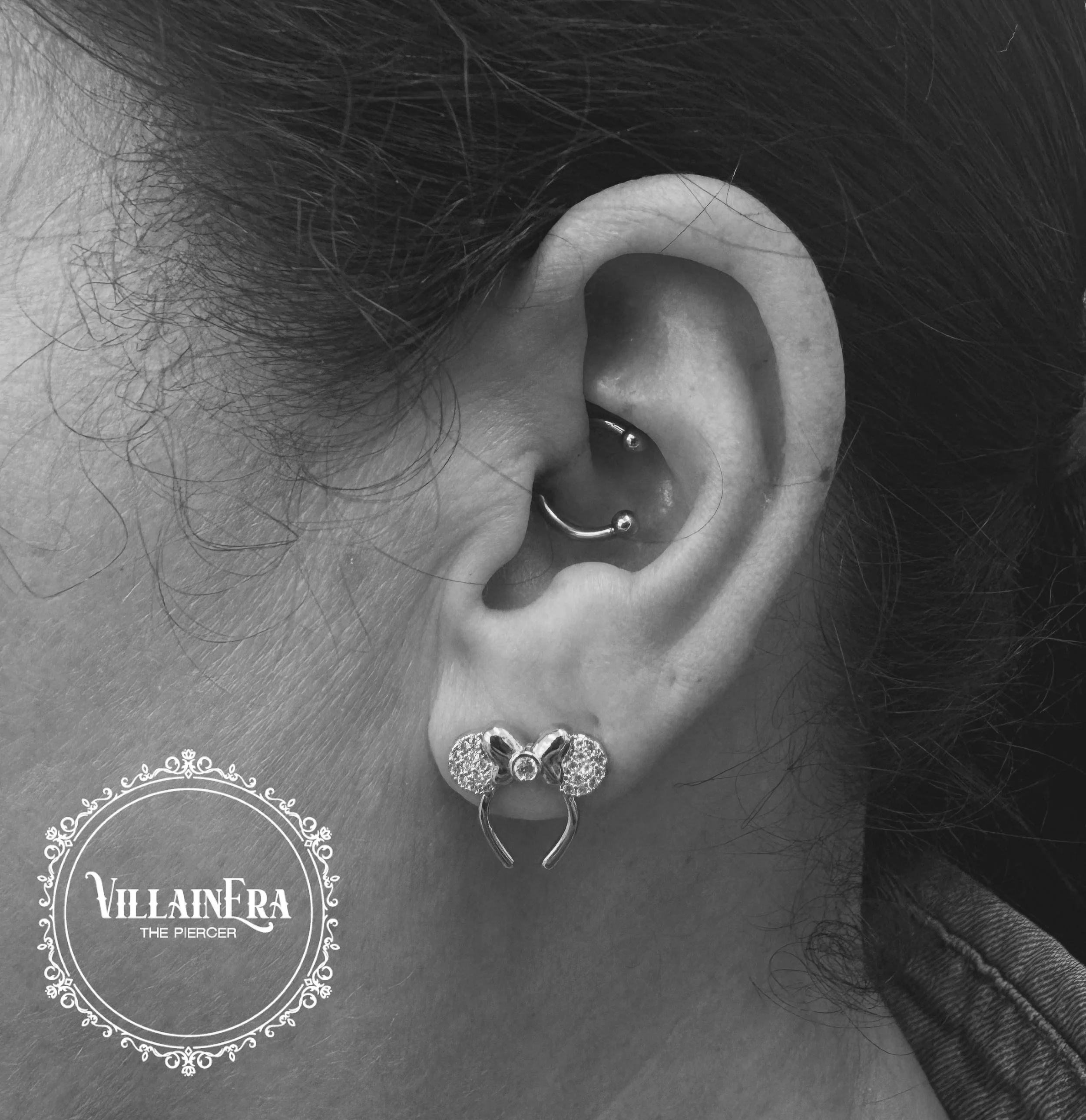 Close-up of a person's ear with multiple piercings, including a cartilage ring and a lobe earring, with hair and a collar visible in the background, and the logo 'VillinEra The Piercer' in the lower left corner.