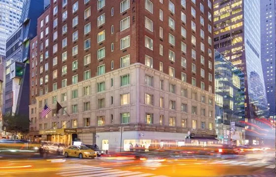 City street with a tall multi-story brick and stone building, yellow taxis, and streaks of lights from moving vehicles at night.