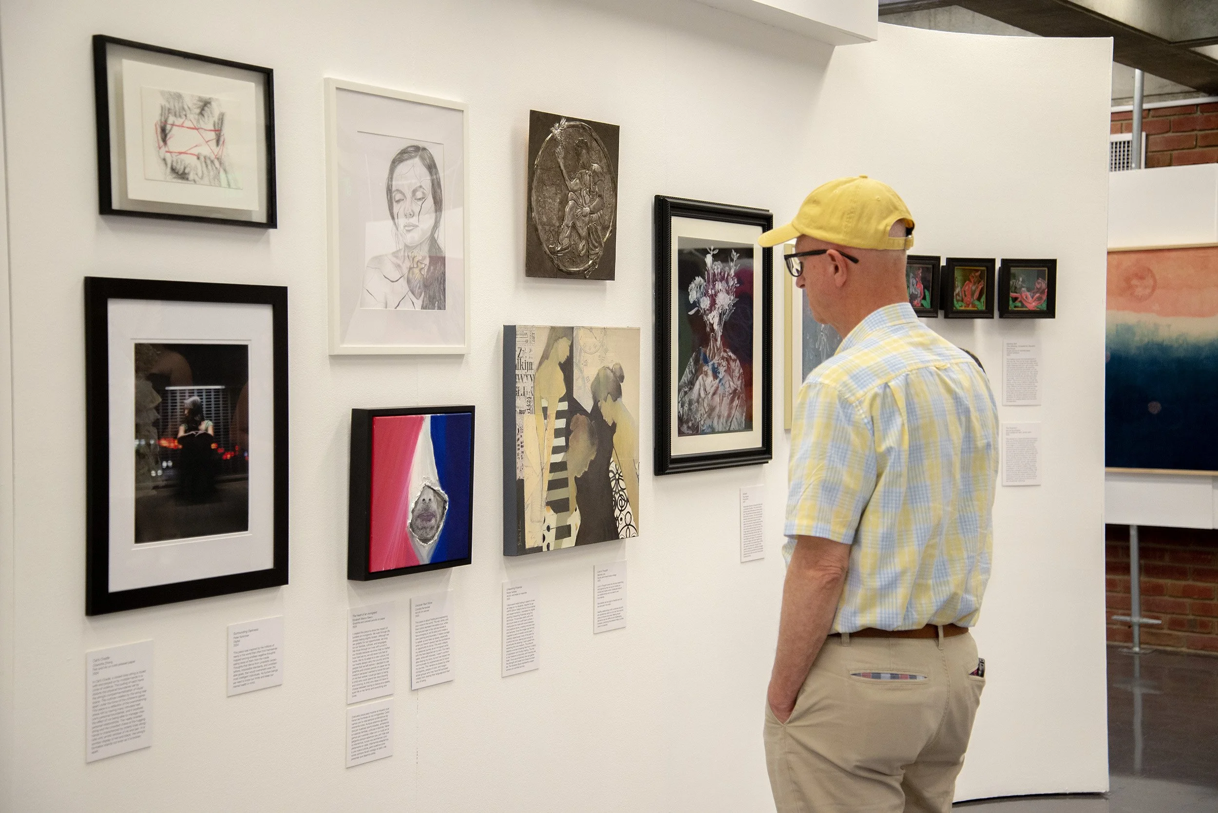 A man wearing a yellow cap, glasses, and a light plaid shirt observes artwork displayed on a gallery wall. The gallery wall features various framed paintings and illustrations, including portraits, abstract art, and mixed media pieces.