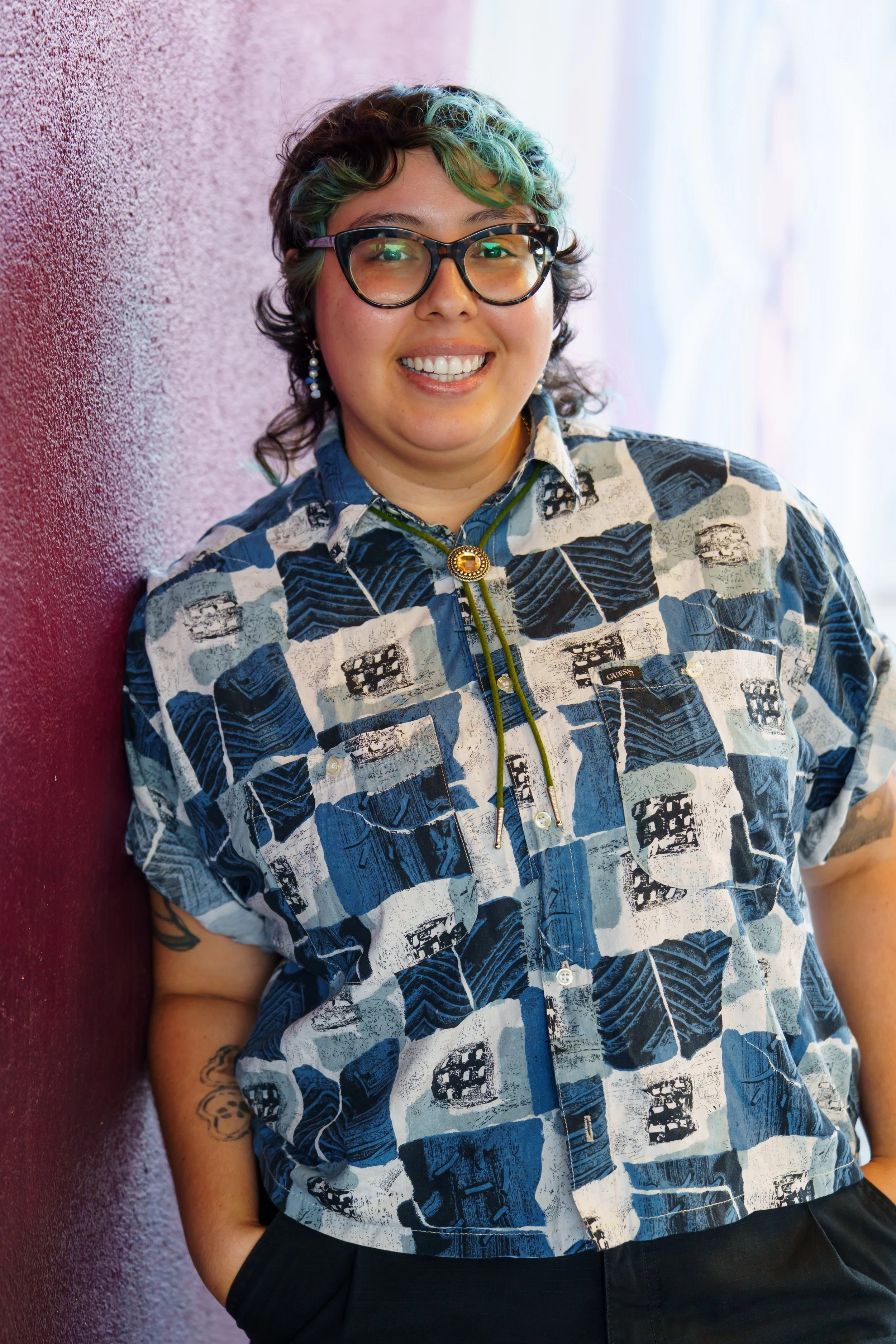A person with glasses and short wavy hair with green highlights, smiling and leaning against a pink wall.