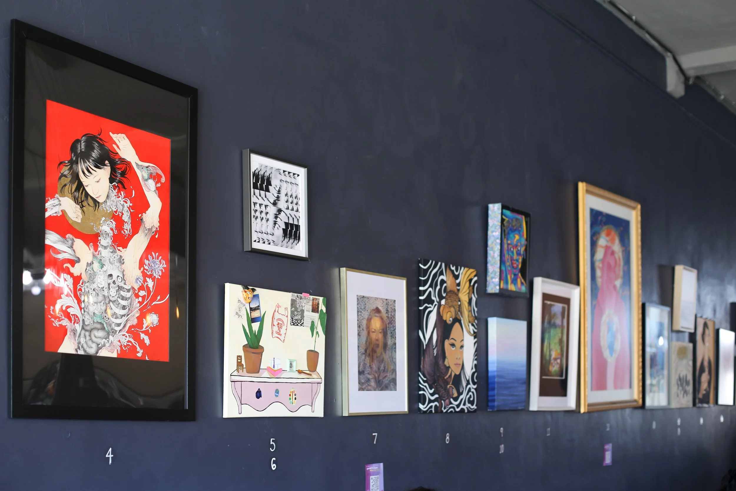Gallery wall with various framed artworks, including a portrait of a woman with long hair, abstract designs, and landscape paintings on a dark blue wall.