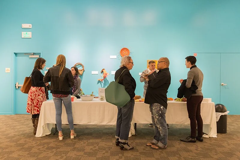 People gathered around a long table with snacks at an indoor event, against a turquoise wall.