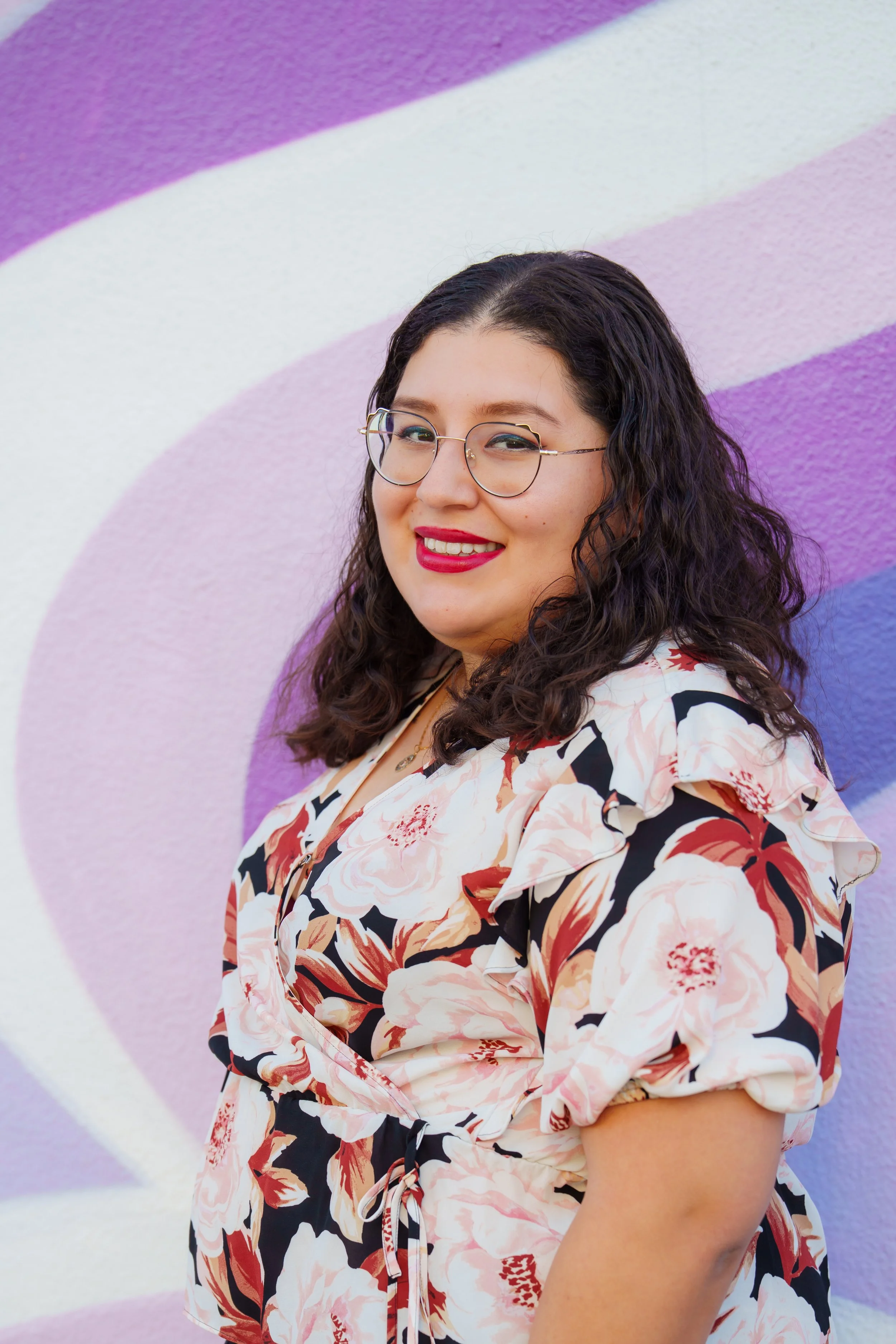 A woman with curly dark hair and glasses, smiling, in front of a colorful mural with purple, pink, and blue shapes. She is wearing a floral blouse.
