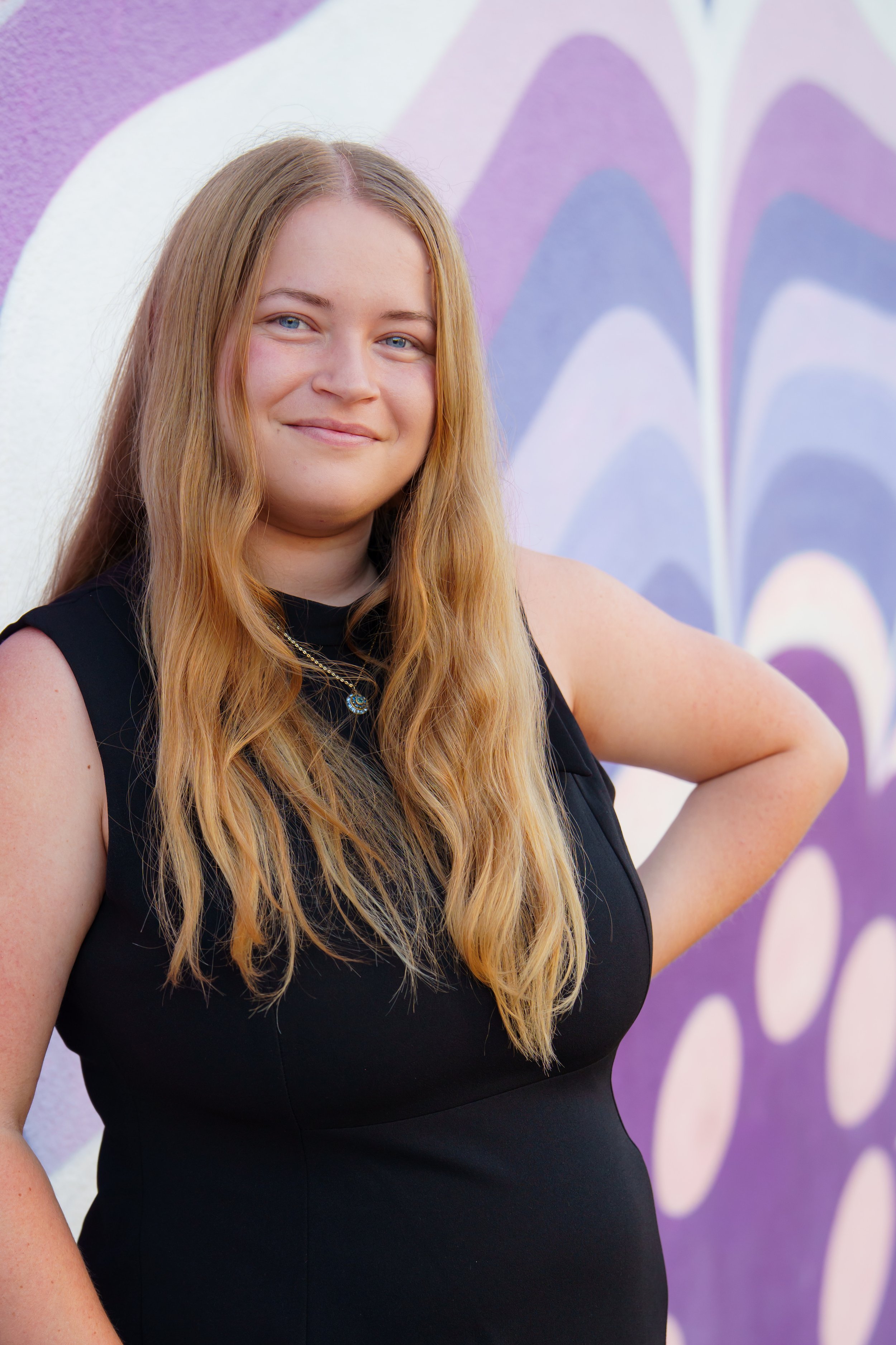 A young woman with long blonde hair and blue eyes standing in front of a purple and white abstract mural, smiling and wearing a sleeveless black top, with one hand on her hip.