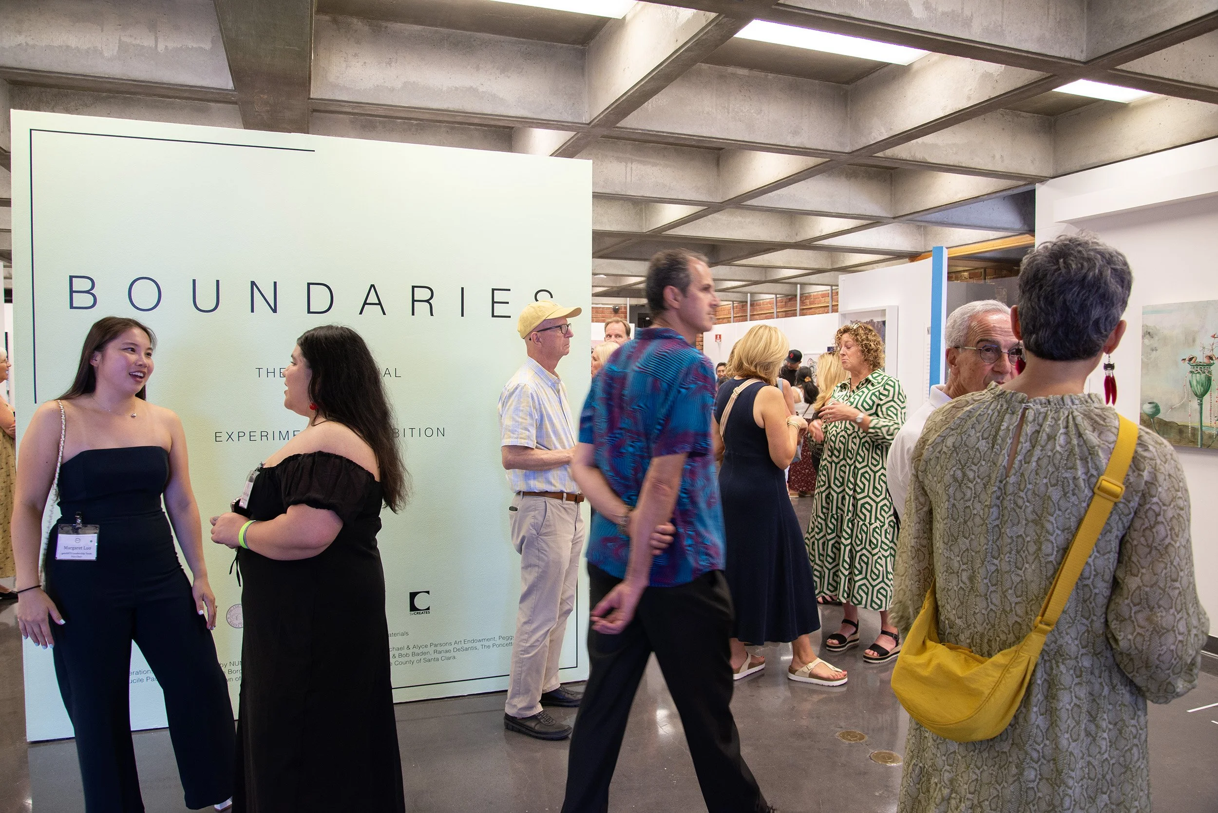An art exhibition with various people viewing artwork and engaging in conversation, with a large sign reading 'Boundaries' in the background.