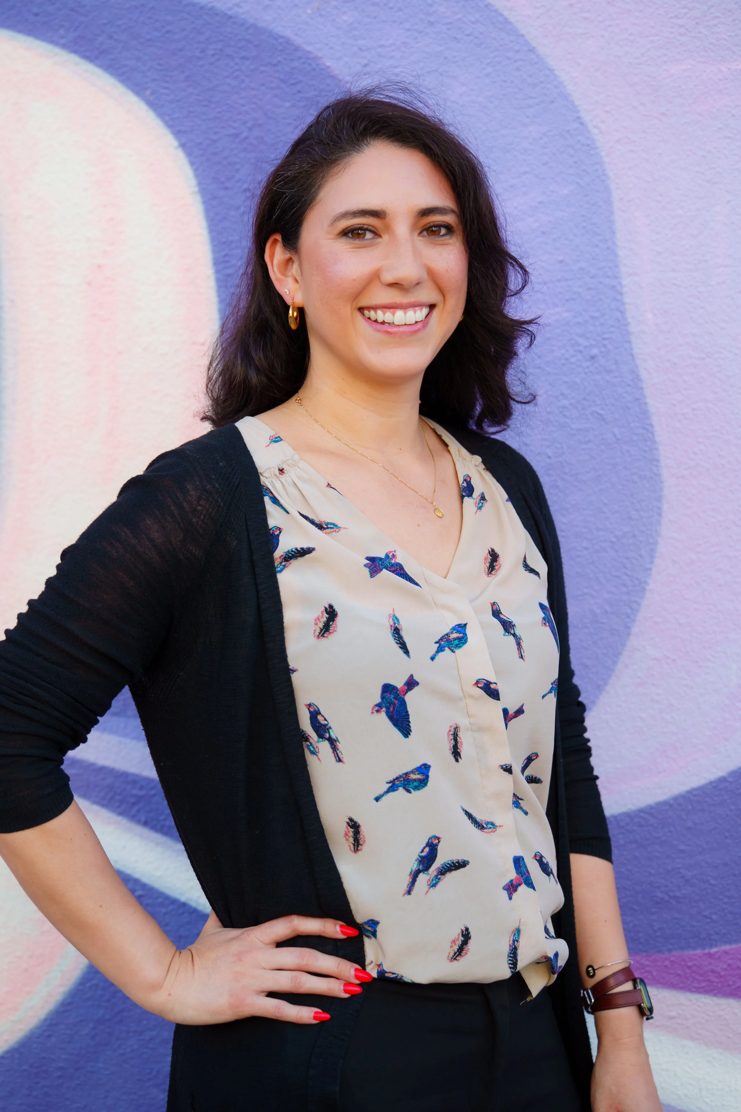 A woman with dark, curly hair wearing a cream blouse with blue bird patterns, a black cardigan, and red nail polish, standing in front of a colorful abstract mural.