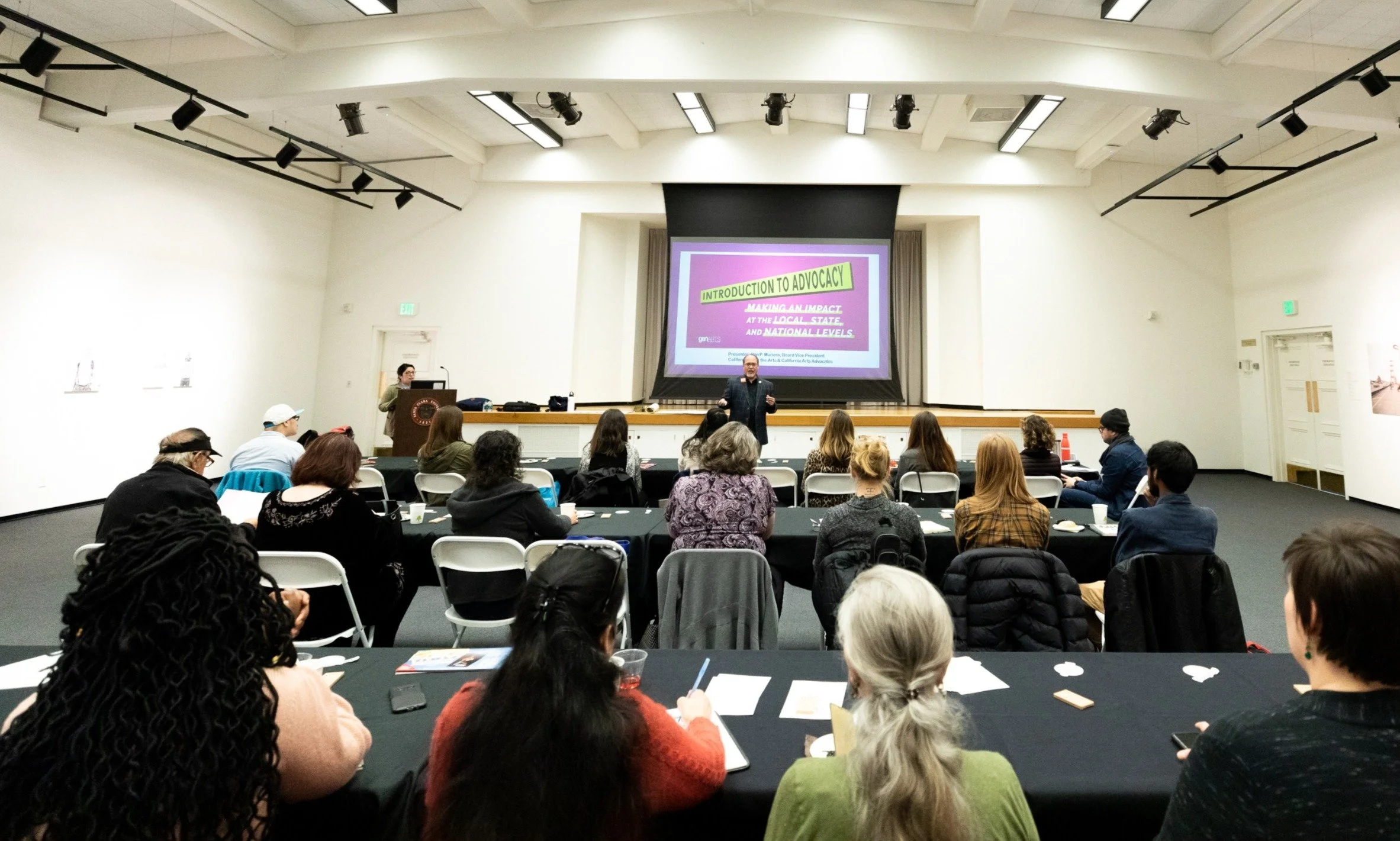 A presentation in a large conference room with a speaker at the front and a group of attendees seated at tables, watching a slide titled 'Introduction to Advocacy' on a large screen.