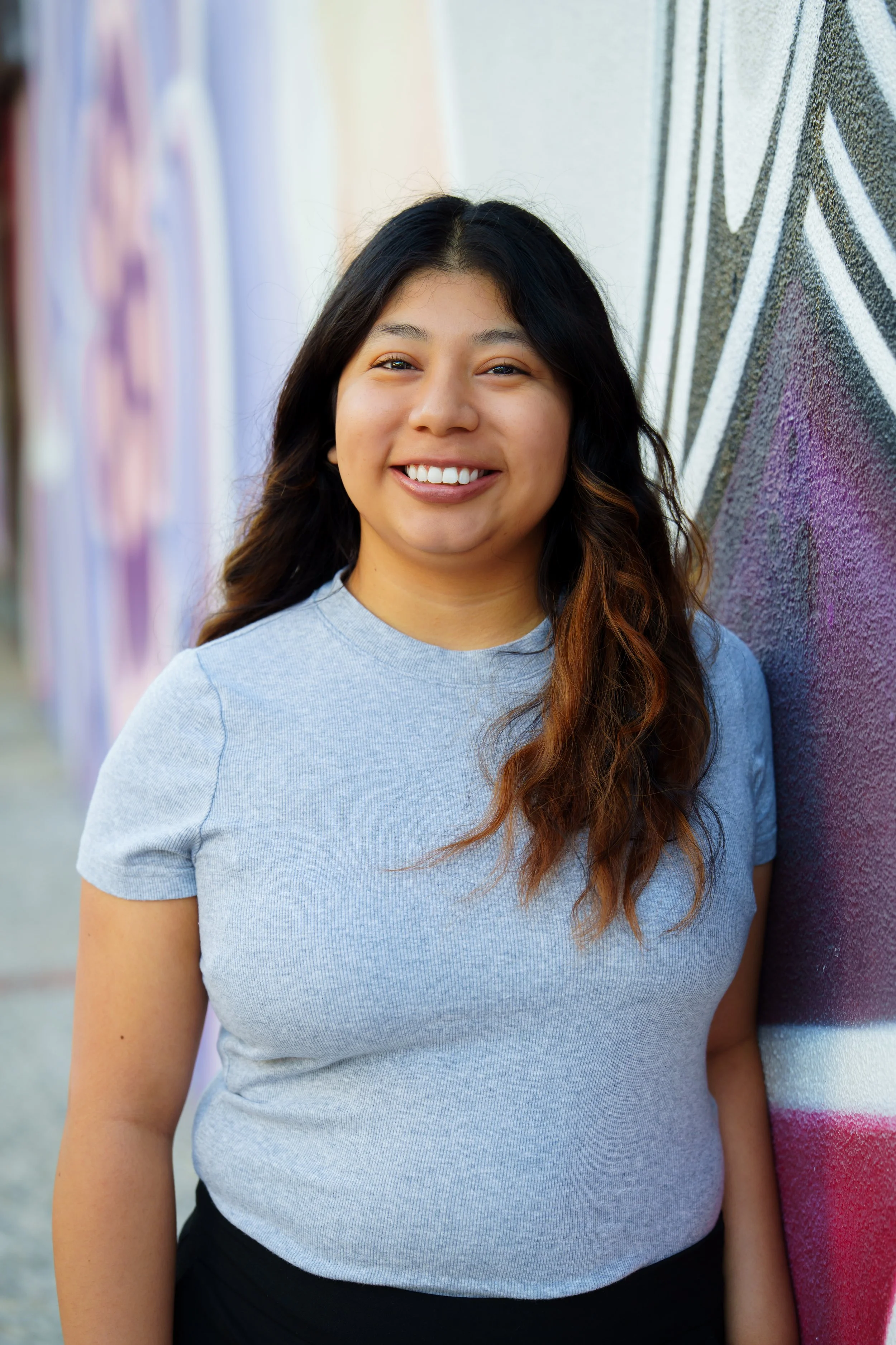 A smiling woman with long wavy hair, wearing a light gray t-shirt, standing next to a colorful graffiti wall.