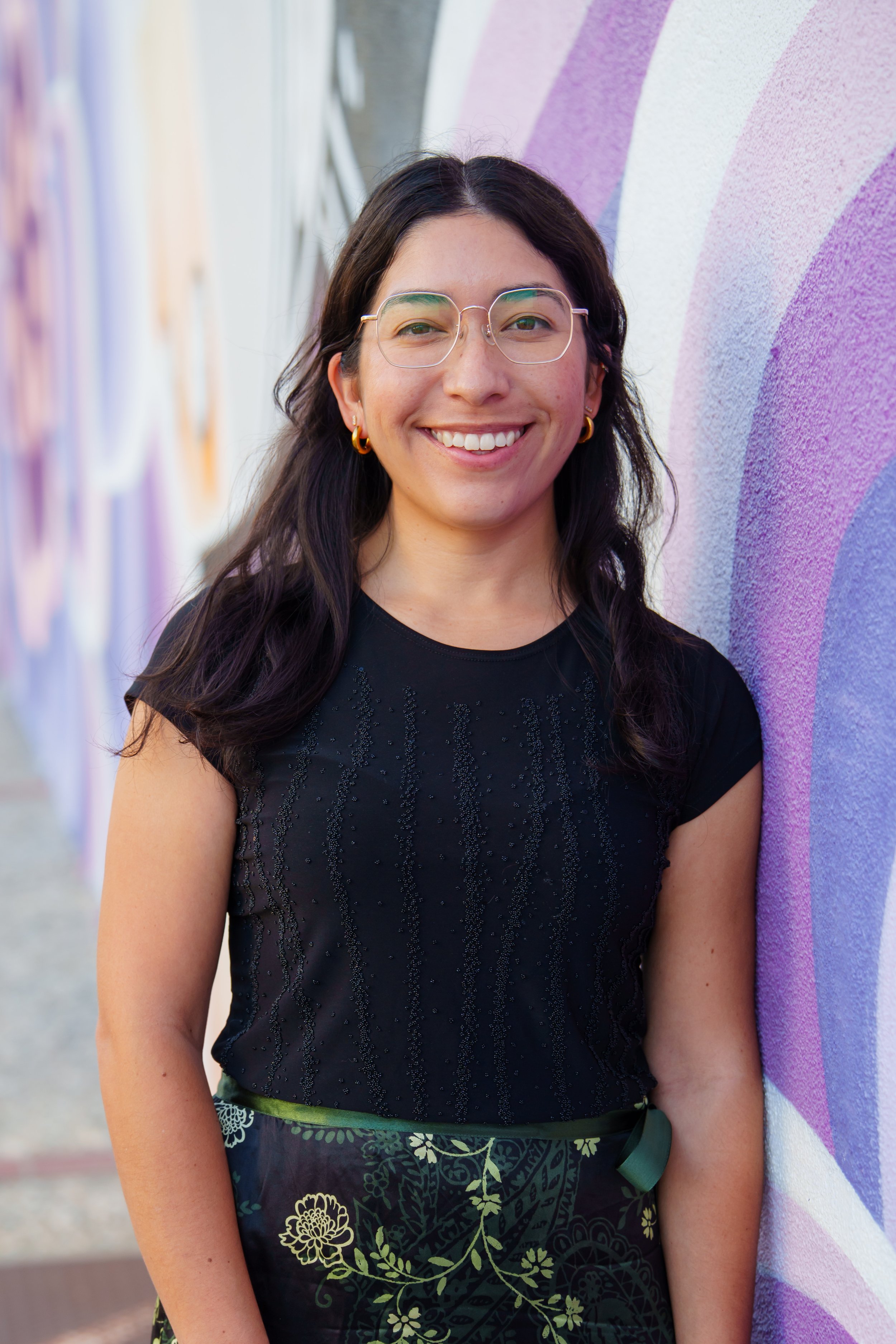 A woman with glasses and earrings smiling while standing against a colorful, purple and pink abstract mural wall.
