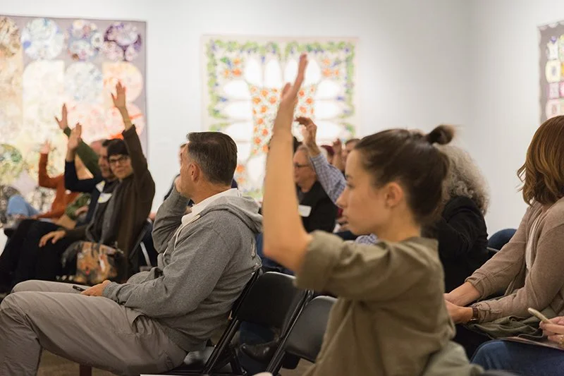People attending a meeting or seminar, raising their hands, seated in chairs in a room with colorful artwork on the walls.