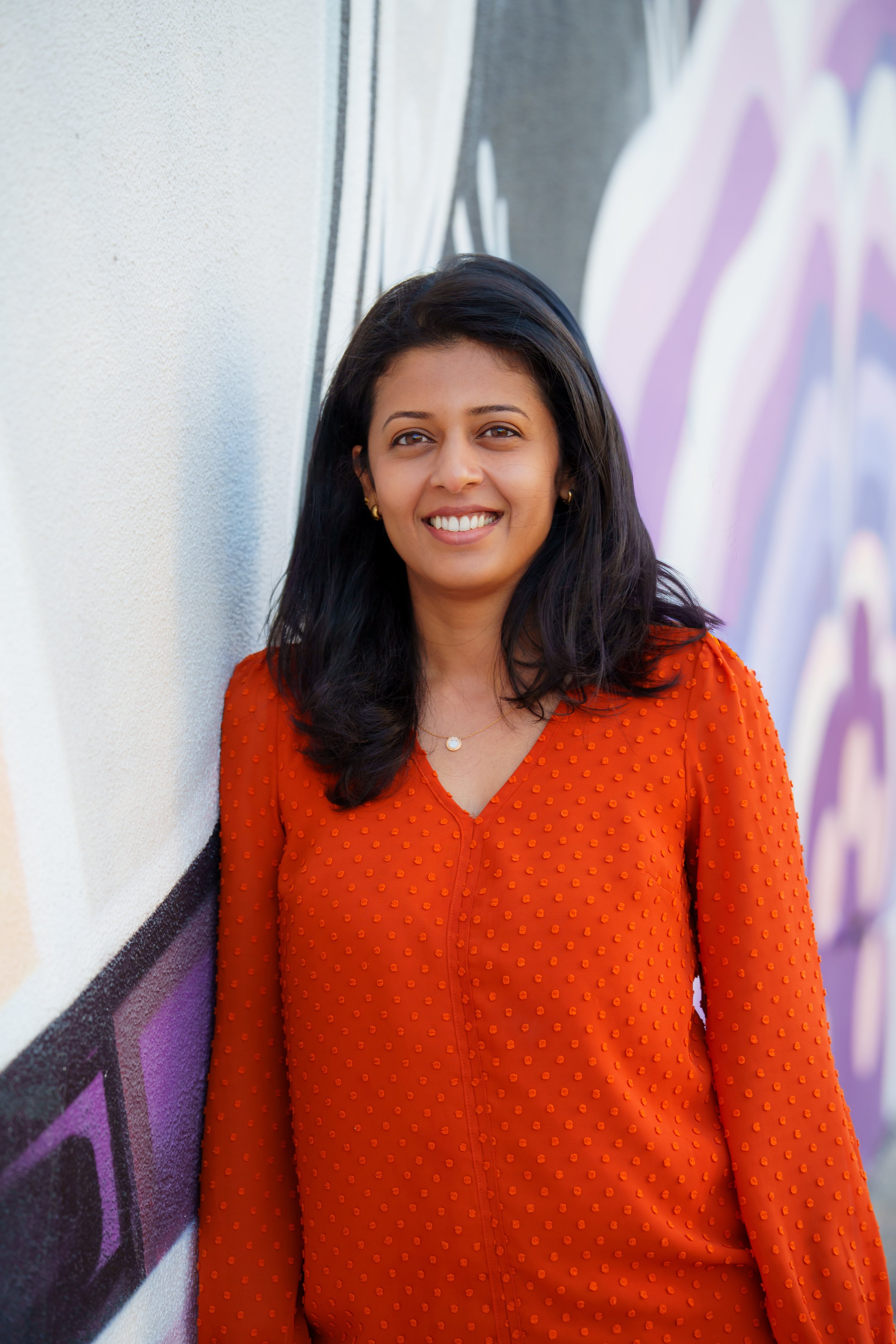 A woman with black hair, wearing a bright orange dress with a textured pattern, standing against a colorful graffiti mural wall, smiling at the camera.