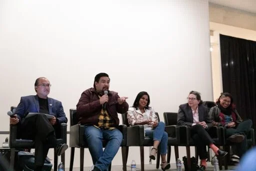 A panel of five people sitting on a stage with black chairs, participating in a discussion or presentation.