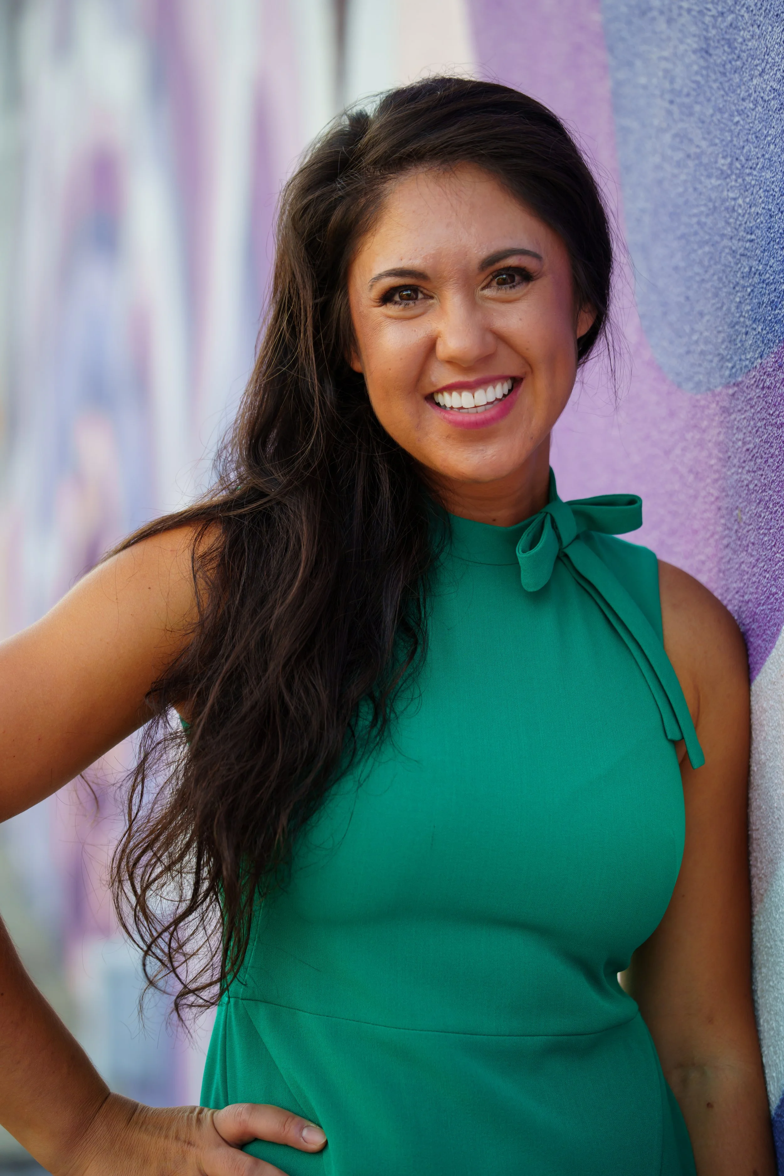 A smiling woman with long dark hair wearing a sleeveless green top with a bow at the neck, standing against a colorful graffiti wall.