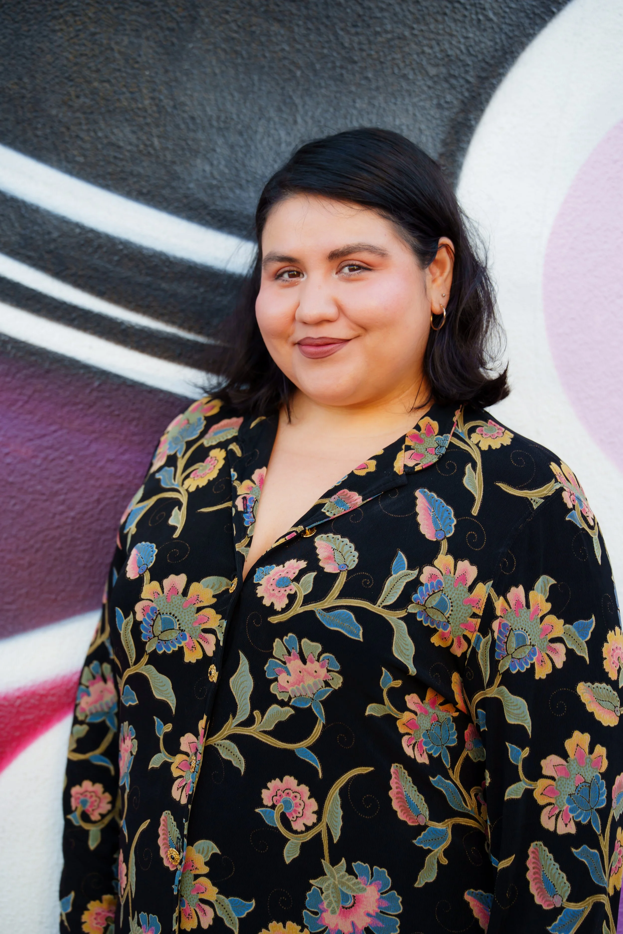 A young woman with black hair and earrings standing in front of a colorful graffiti wall, smiling slightly, wearing a black floral patterned top.