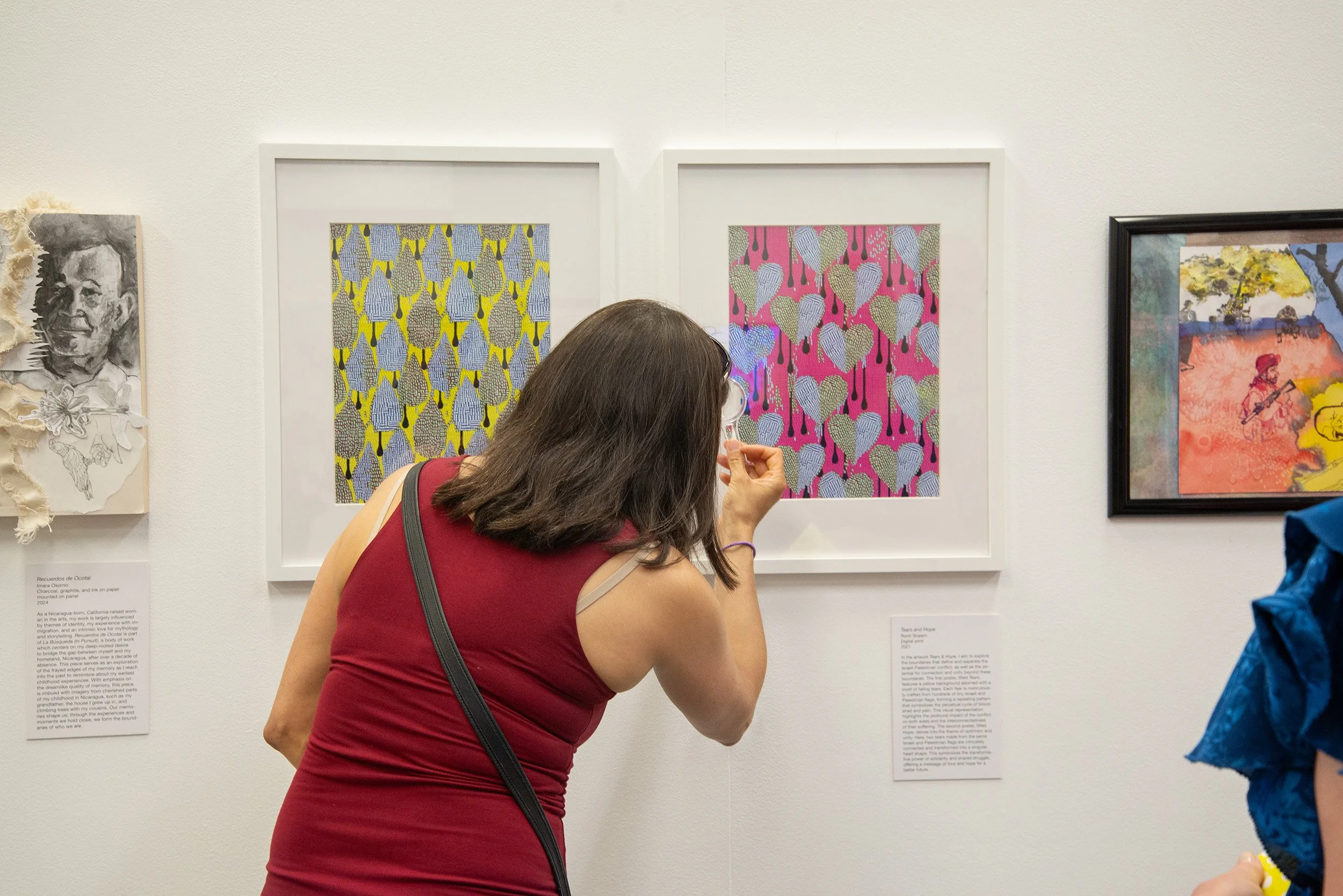 A woman with brown hair, wearing a red sleeveless dress and glasses, is looking at and photographing two framed abstract artworks with a smartphone in an art gallery.
