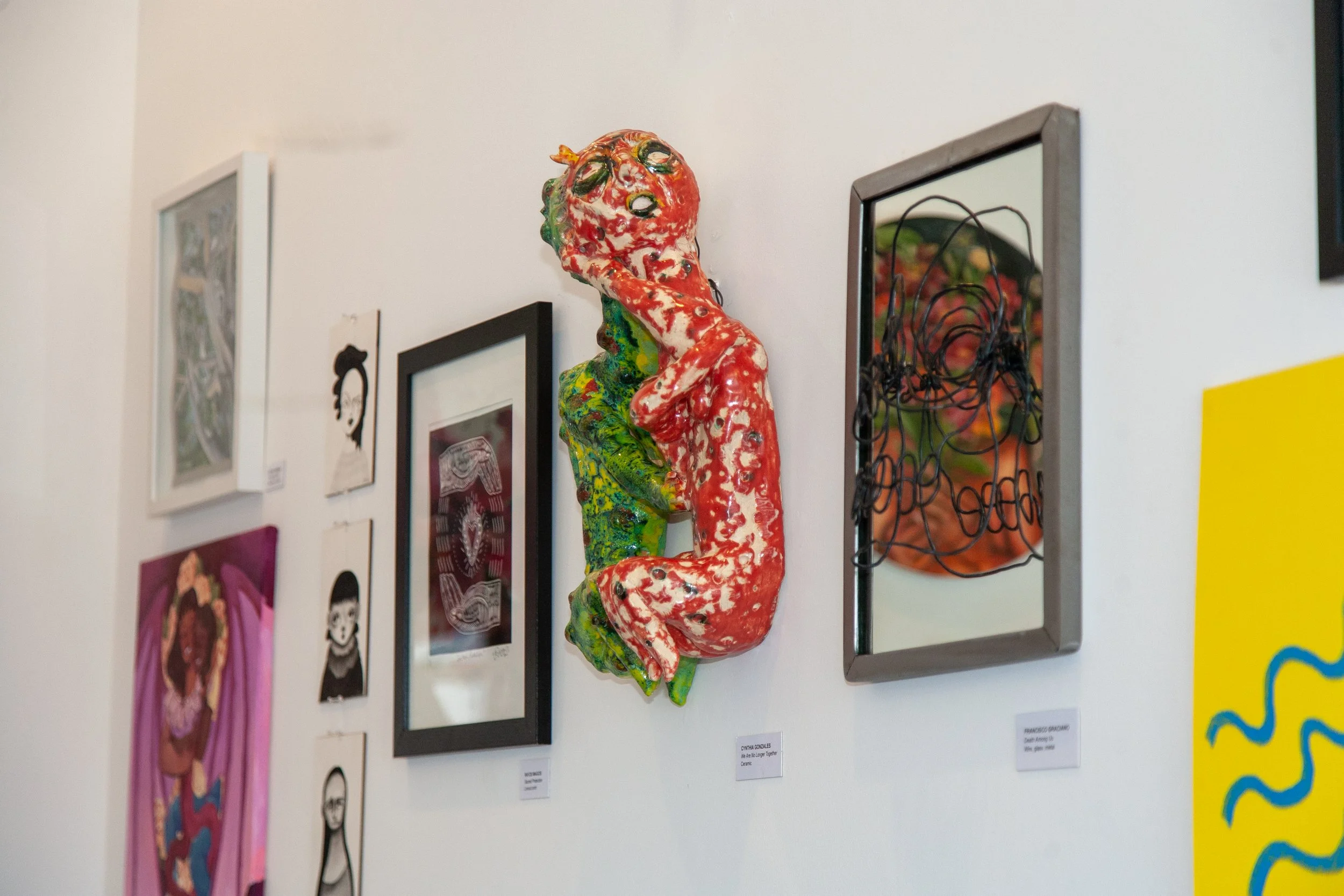 Colorful ceramic sculpture of a curled-up cat with a glossy, textured surface hanging on a white gallery wall, surrounded by framed artwork.