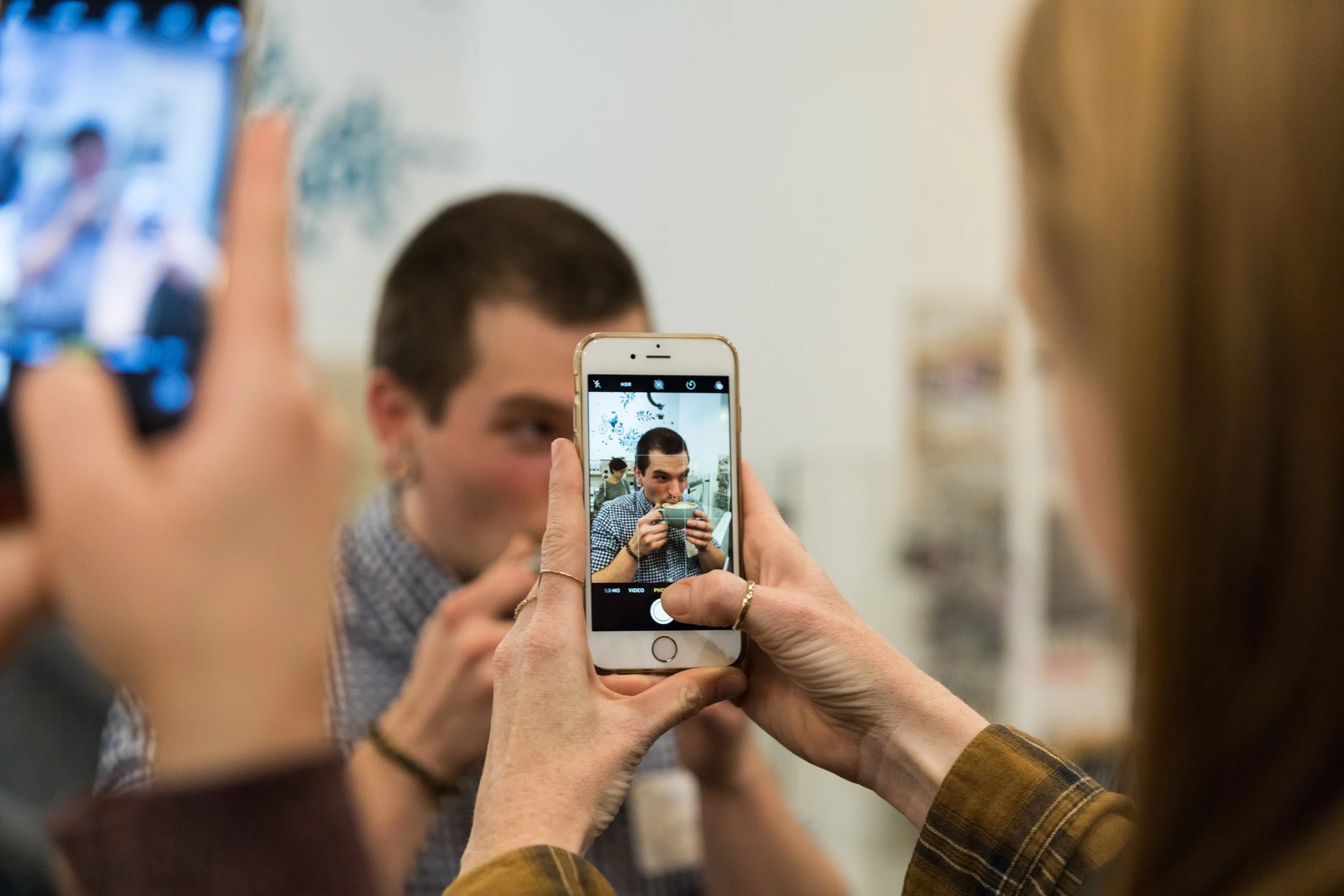 Person holding a smartphone taking a photo of a young man sipping from a mug.