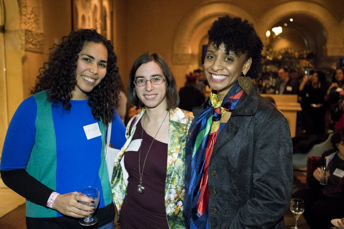 Three women smiling at a social event in a decorated hall. The woman on the left has curly hair, wears a blue and green shirt, and holds a glass of water. The woman in the middle has straight hair, glasses, and wears a patterned jacket over a burgund