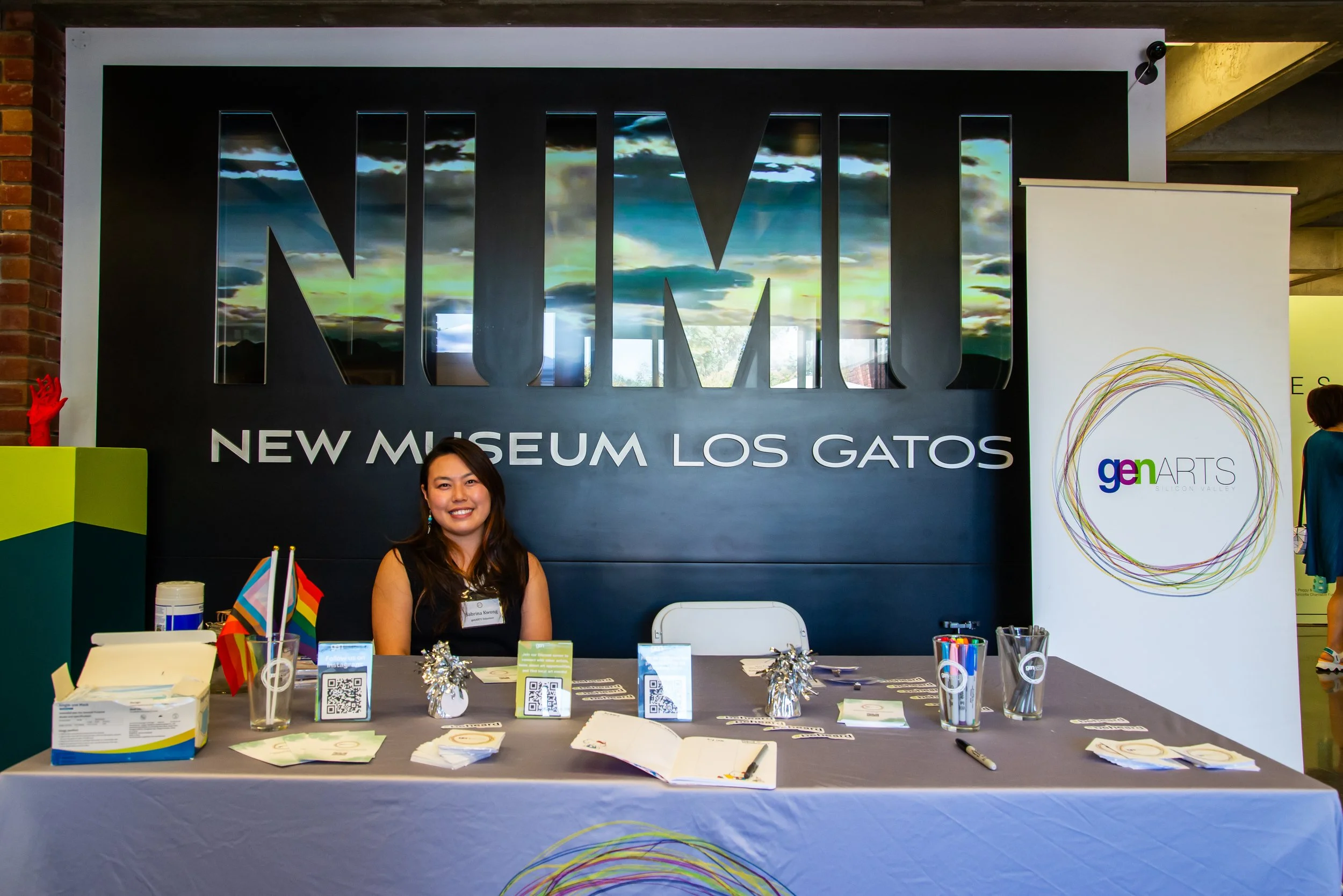 A woman sitting behind a table at a conference or event, with a large sign behind her that reads 'NMWL' and 'New Museum Los Gatos.' The table has various papers, QR codes, and small flags, with a colorful sign to her right.