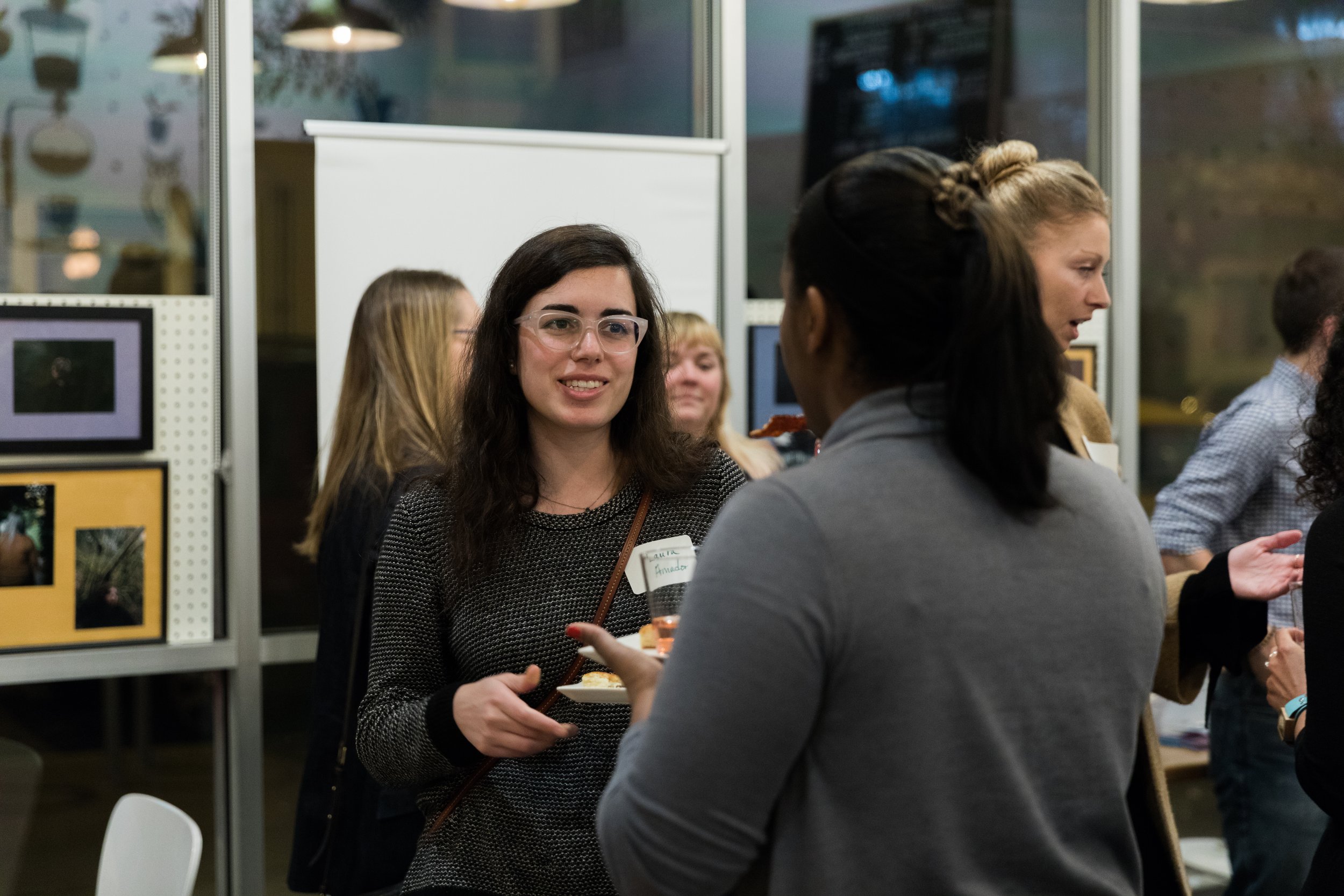 Women engaging in conversation at an indoor networking event, with art or photographs displayed on the walls in the background.