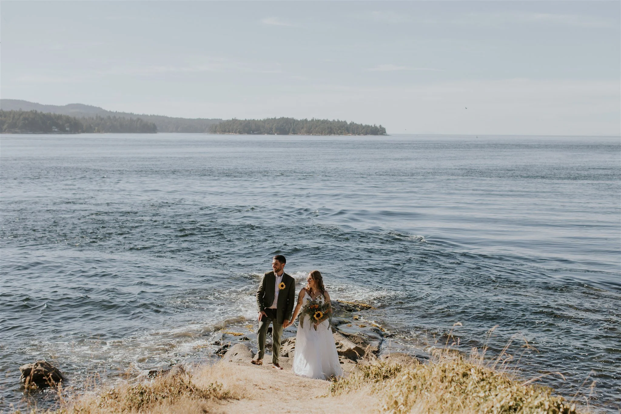 A bride and groom standing hand in hand near the shoreline of a calm body of water with a forested island in the background, on a breezy day.
