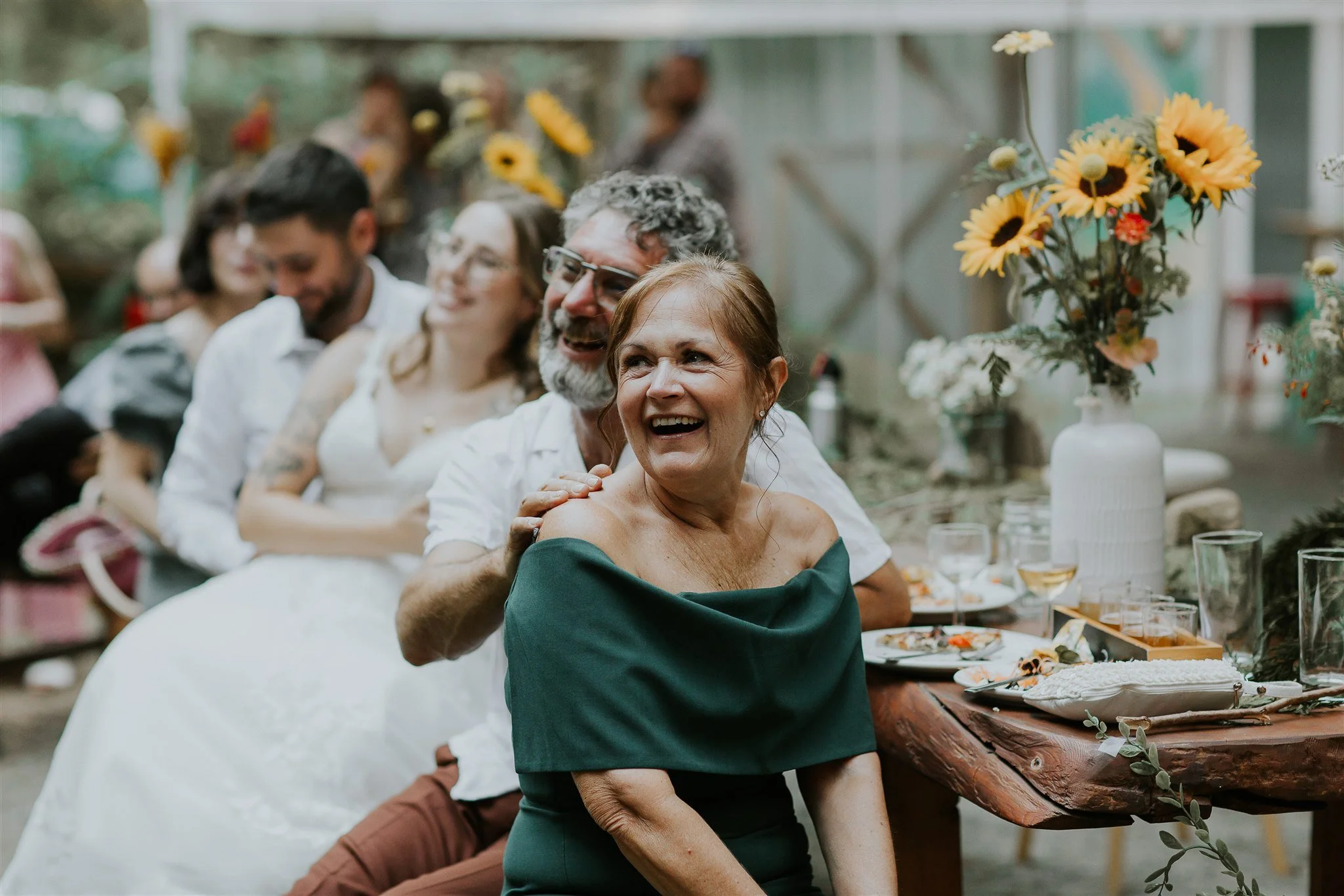 Group of people happily sitting at a table with flowers and food, celebrating at a joyful event, possibly a wedding reception.