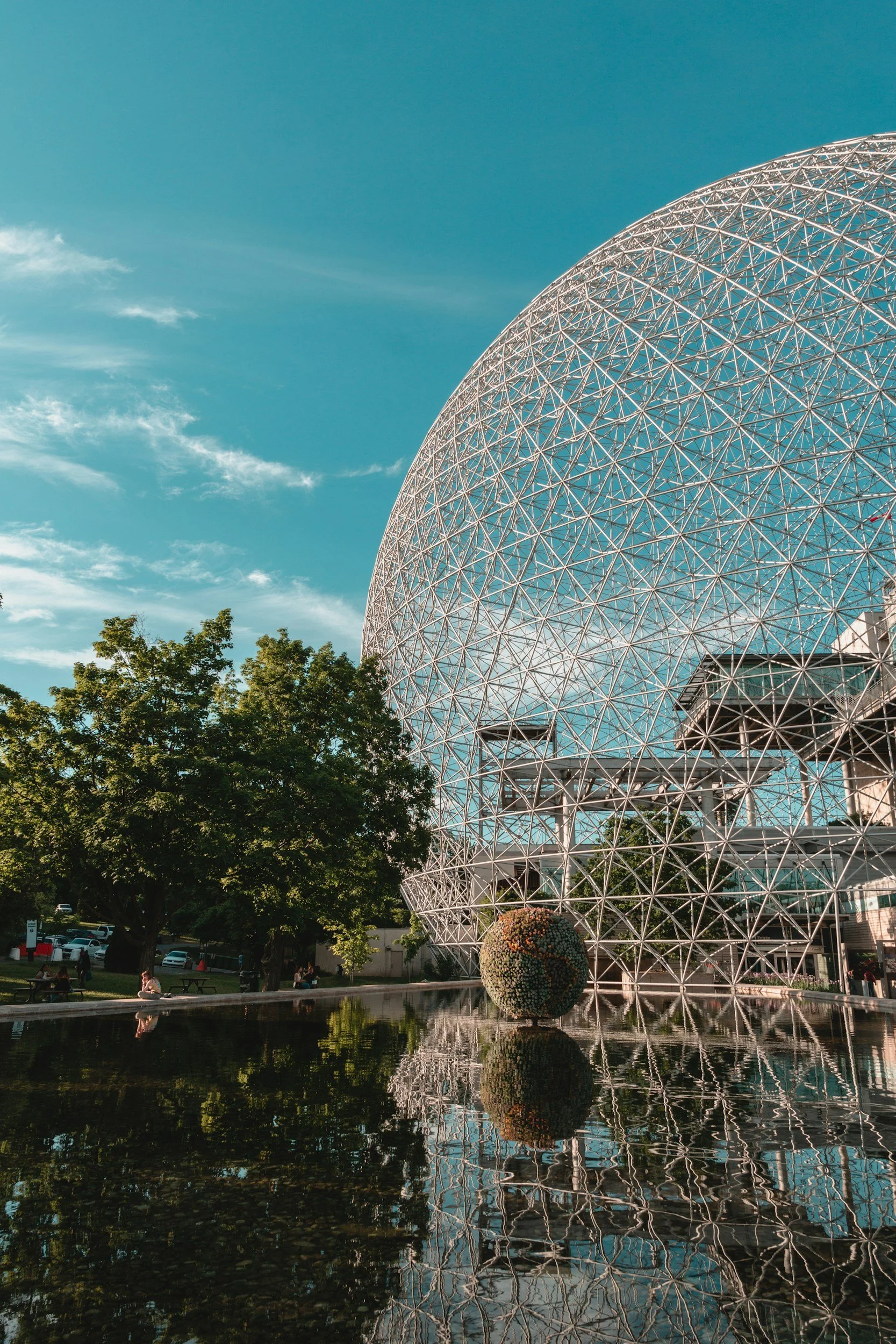 The image features a large spherical glass and steel geodesic dome with a reflection in a small pond in the foreground, surrounded by trees and a park area.