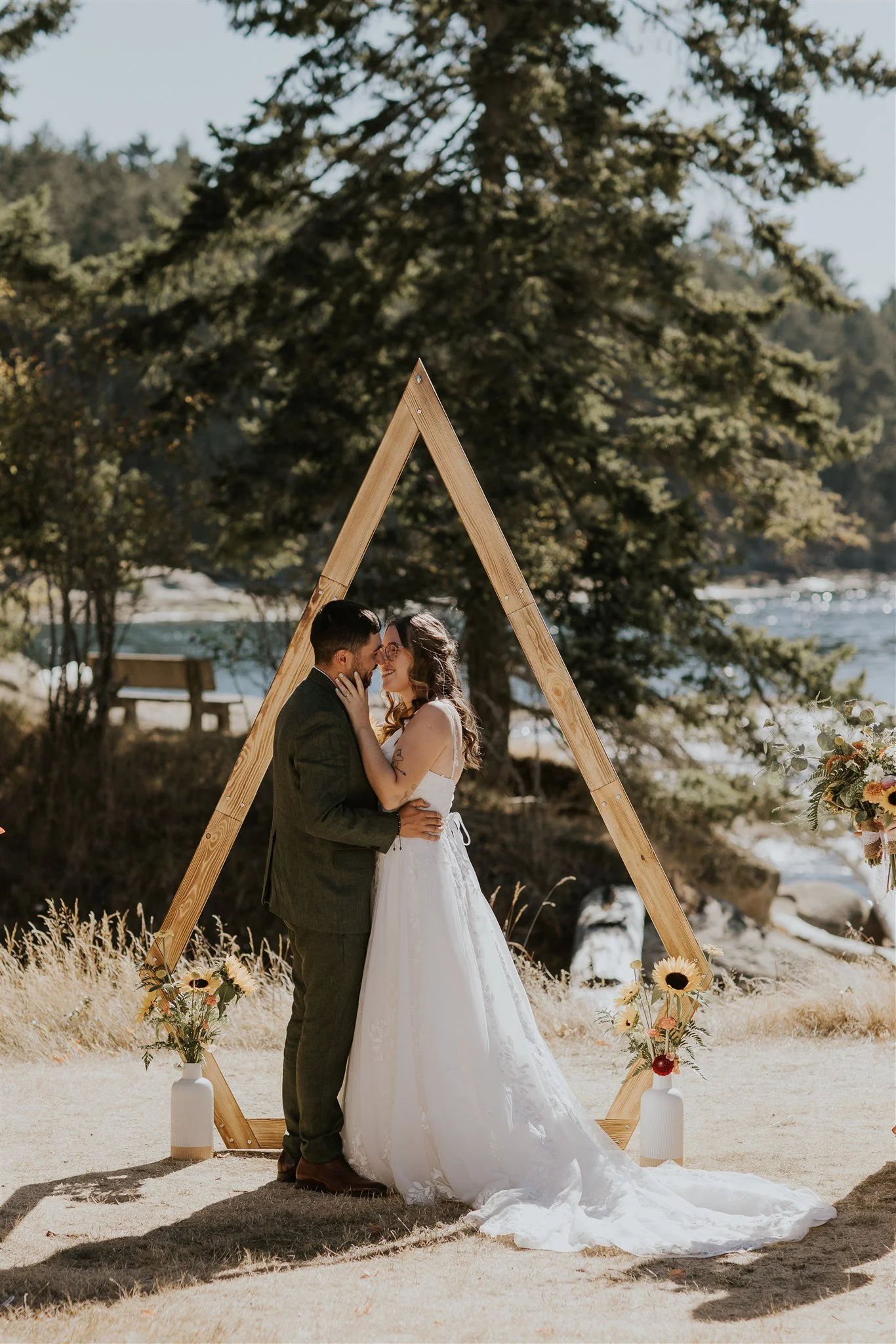 A couple in wedding attire sharing a kiss, standing under a wooden triangle arch decorated with flowers, outdoors near a river with trees in the background.