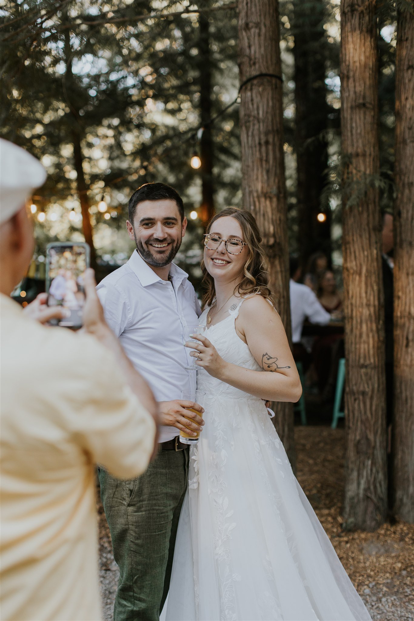 A couple smiling and posing for a photo at a wedding reception outdoors, with trees and string lights in the background, while a person in a yellow shirt takes their picture.